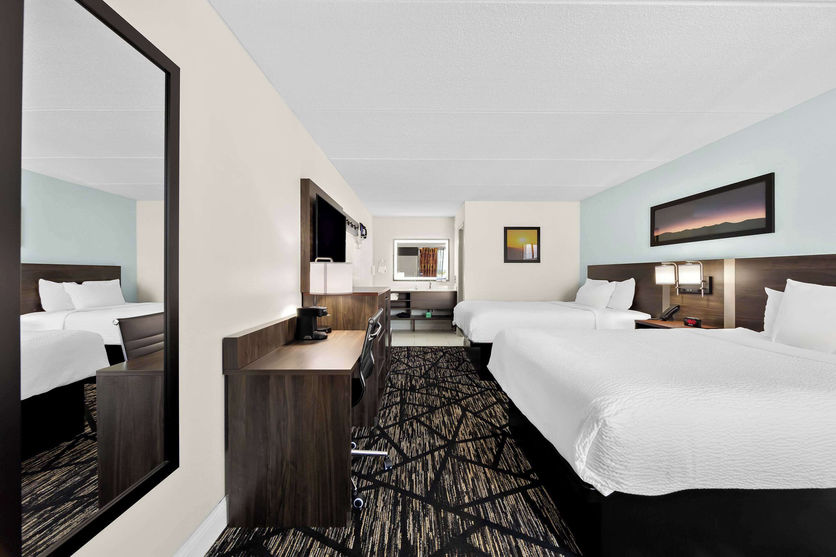 A hotel room with two beds, a desk, a mirror, a TV, and a painting on the wall.