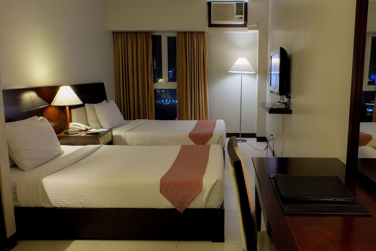 A hotel room with two beds, a desk, a lamp, a TV, and a window with curtains.