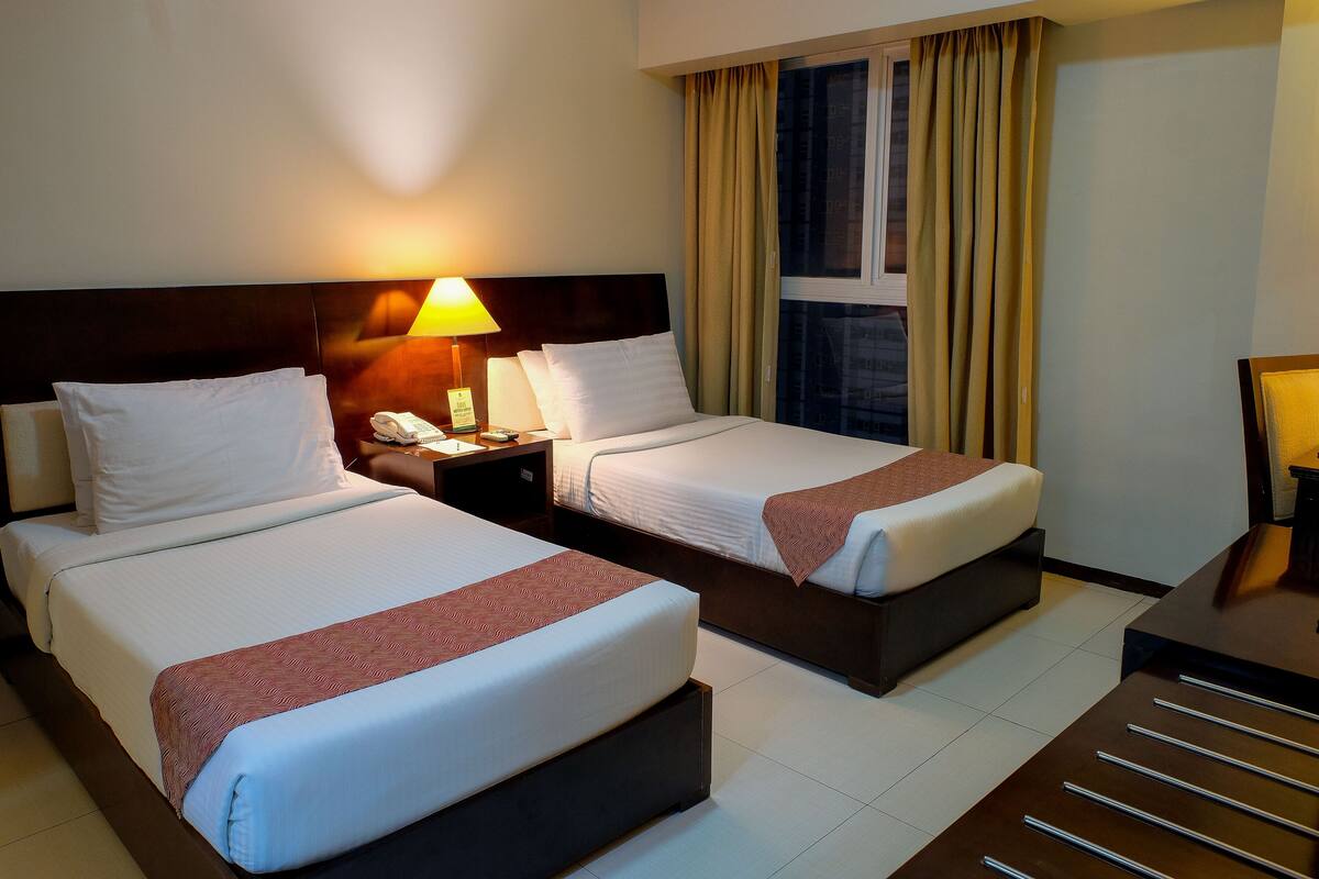A hotel room with two beds, a nightstand with a lamp, a window with curtains, and a desk with drawers.