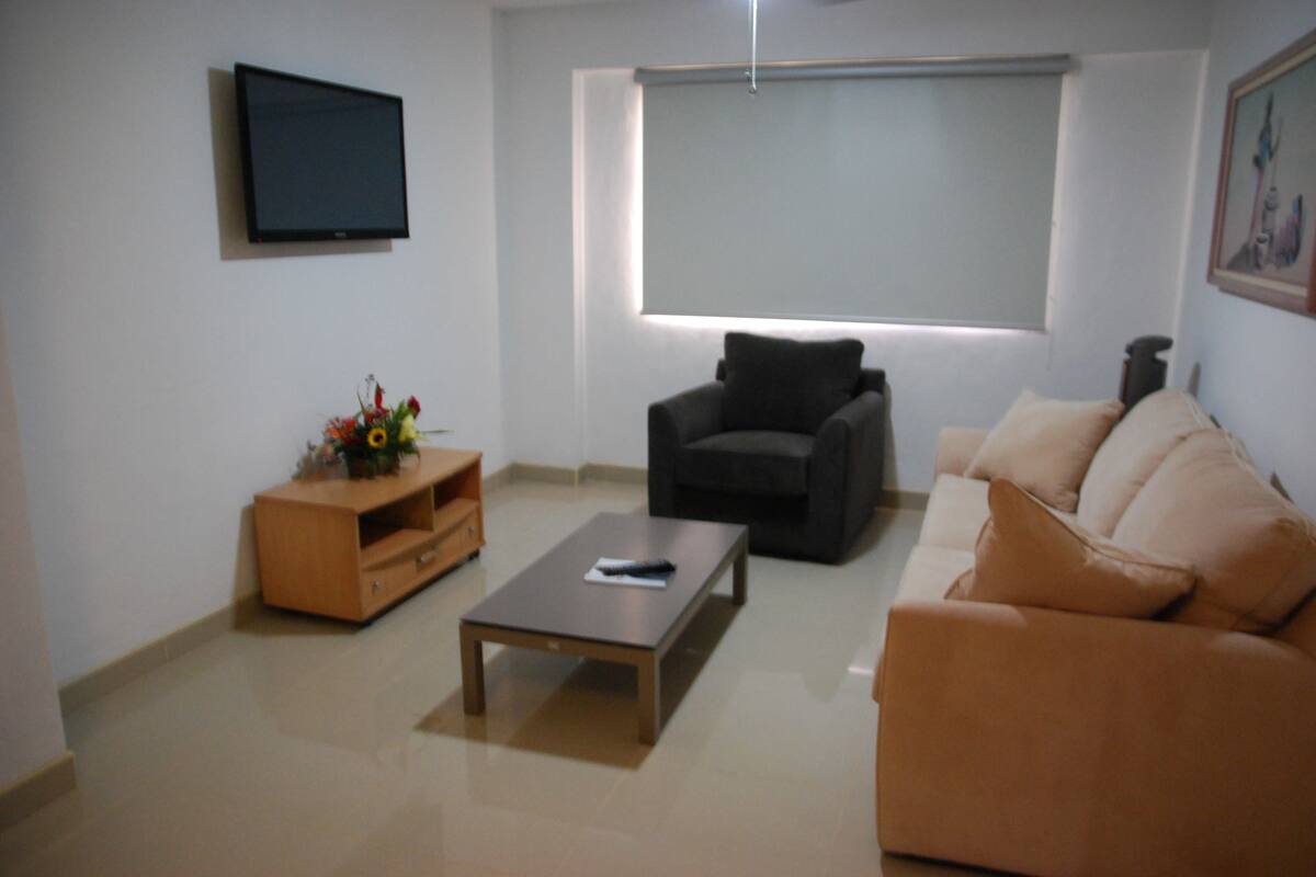 A modern living room with a flat-screen TV, a sofa, a coffee table, and a TV stand with a flower arrangement.