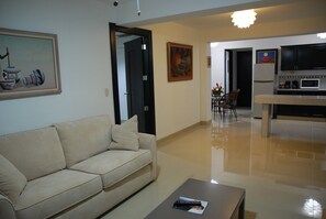 A modern living room with a beige sofa, a coffee table, and a kitchen area with a microwave and cabinets. - Hoteles Brandt (Managua)