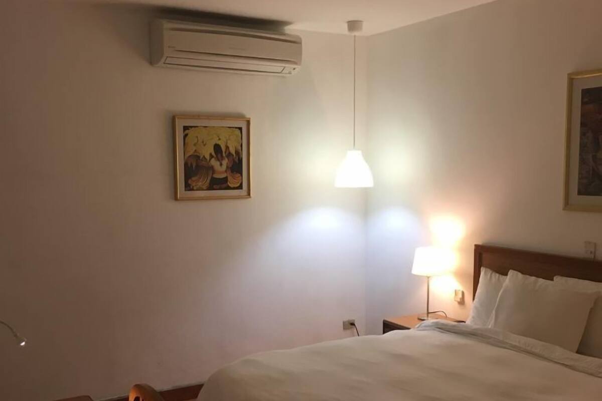 A hotel room with a large bed, a desk, and a chair. There is a painting on the wall and a ceiling fan.