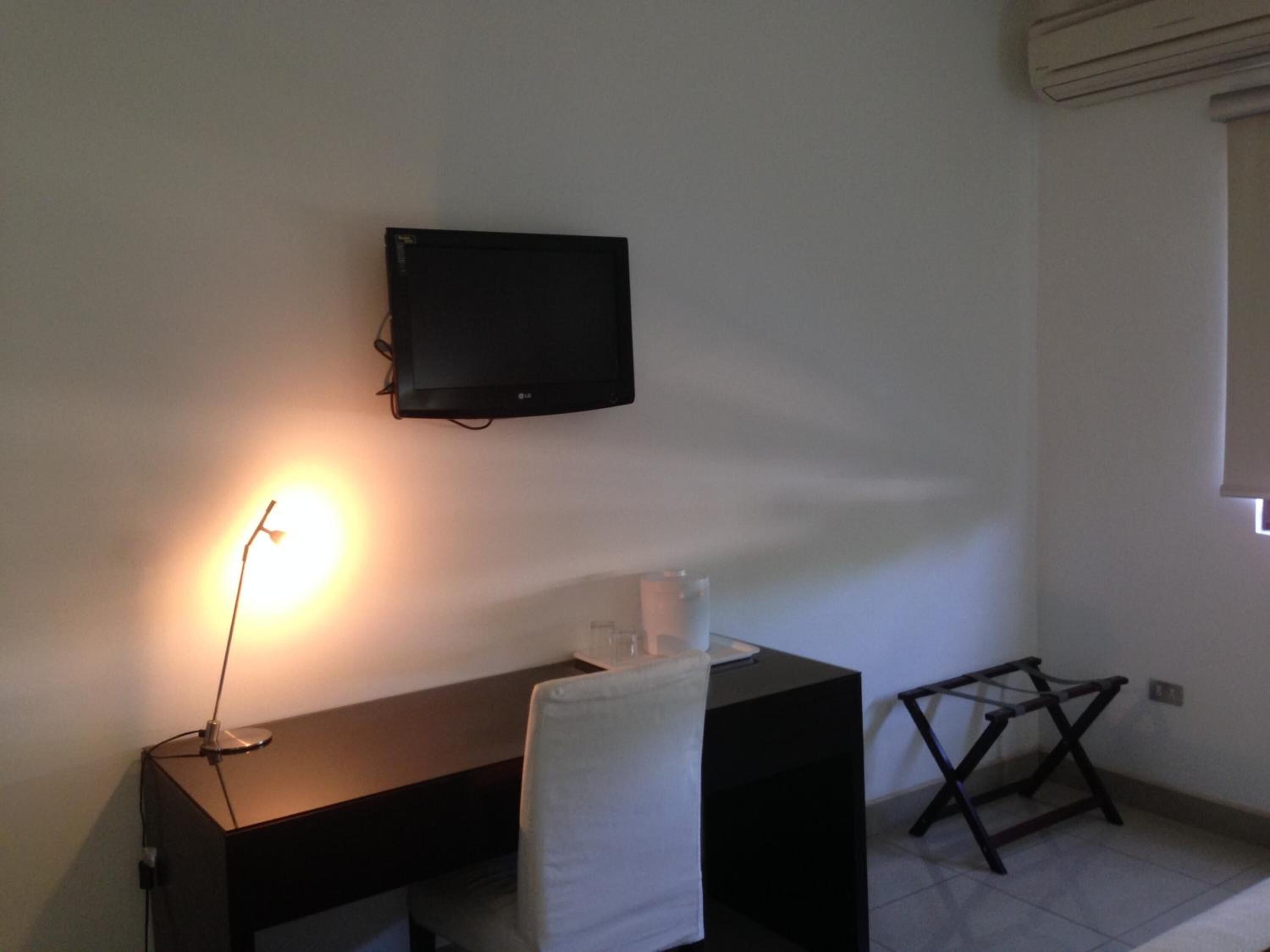 Una habitación pequeña y poco amueblada con un escritorio, una silla, un televisor en la pared y una ventana con persianas.