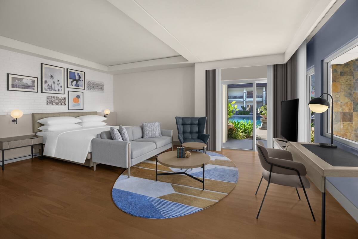 Ein modernes Hotelzimmer mit Bett, Sofa, Schreibtisch und Fernseher. An den Wänden hängen Bilder und es gibt einen Blick auf den Balkon.