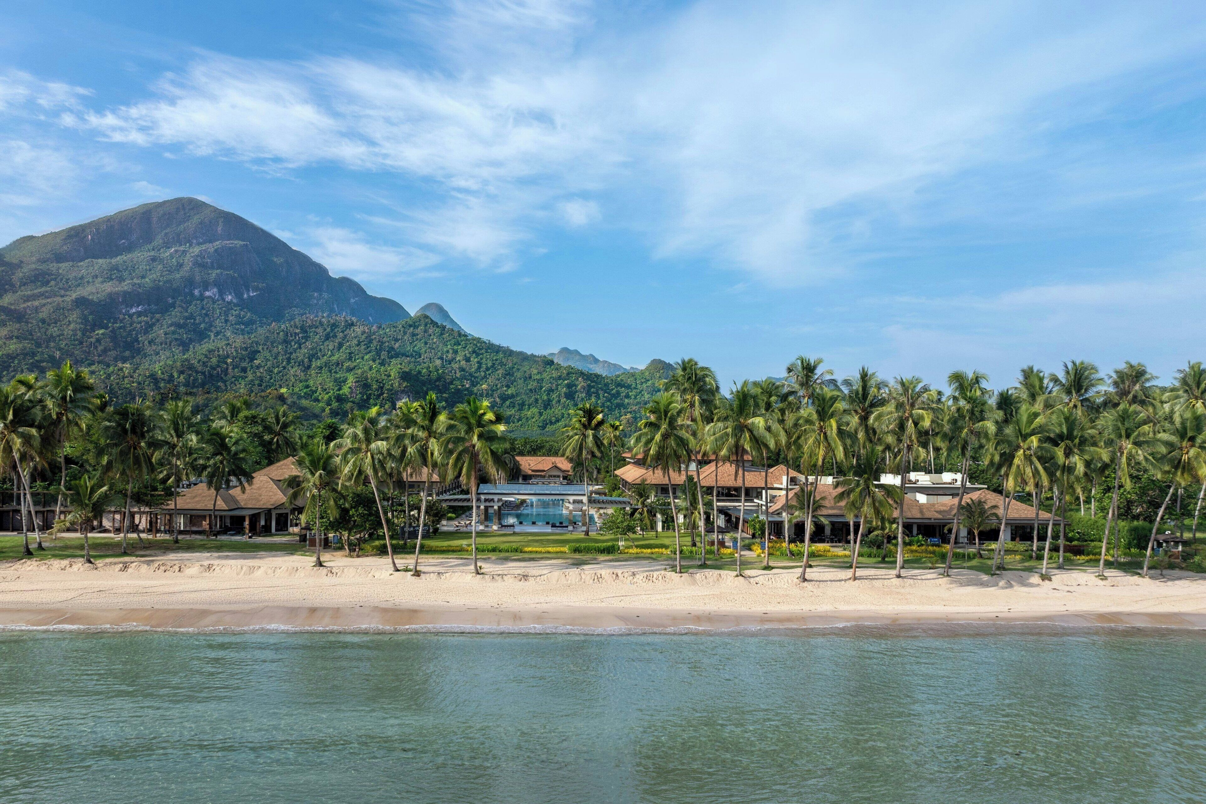 A beachfront property with multiple buildings, palm trees, and a mountain in the background.