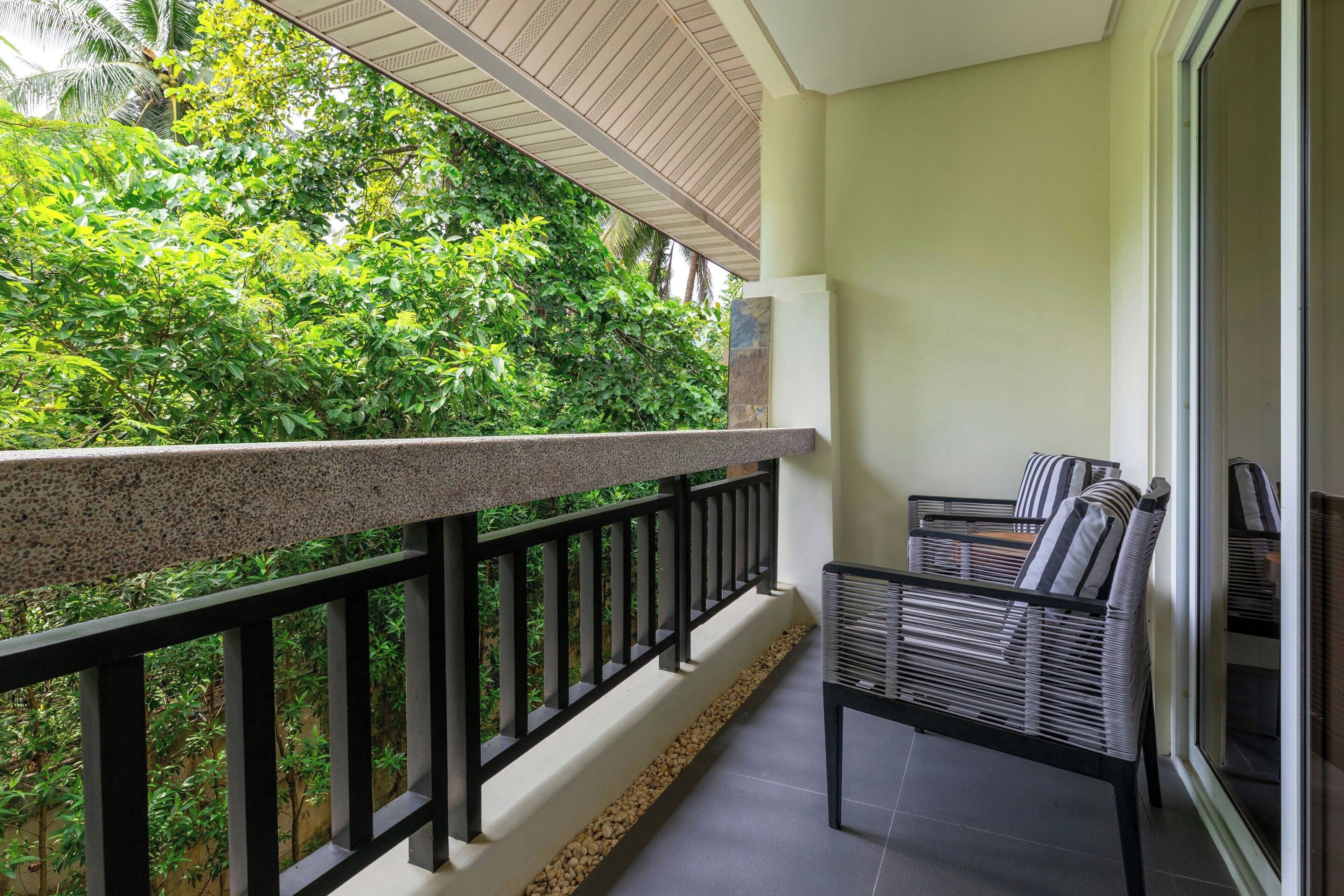 Een balkon met een stenen balustrade, een zithoek met gestreepte kussens, en uitzicht op weelderige begroeiing.