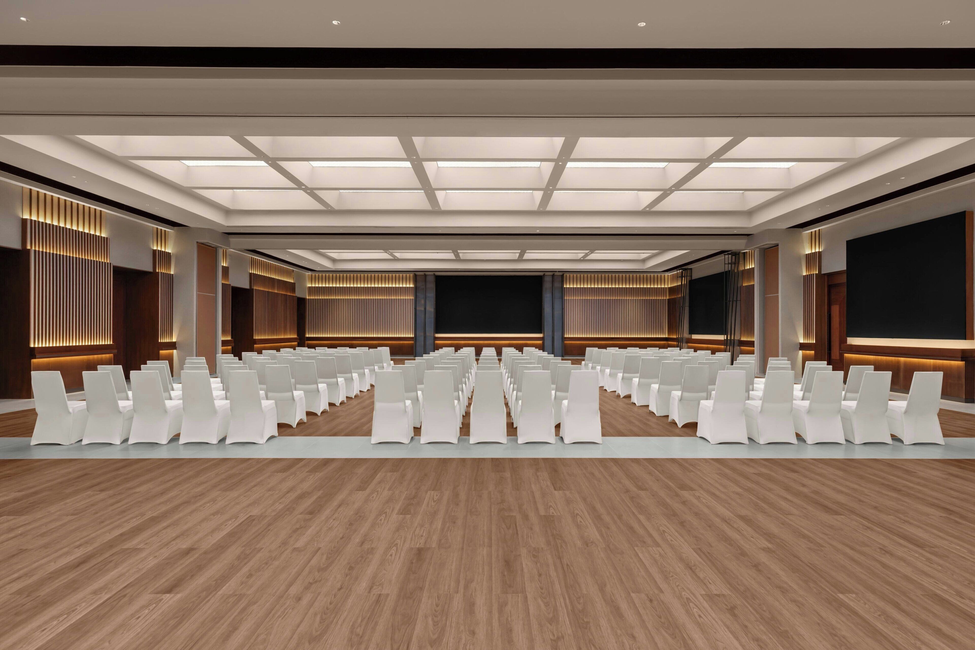 Een vergaderzaal met rijen witte stoelen, een houten vloer en een plafond met inbouwverlichting.