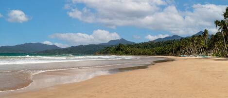 A beach with a clear sky, ocean waves, and a mountain range in the background.