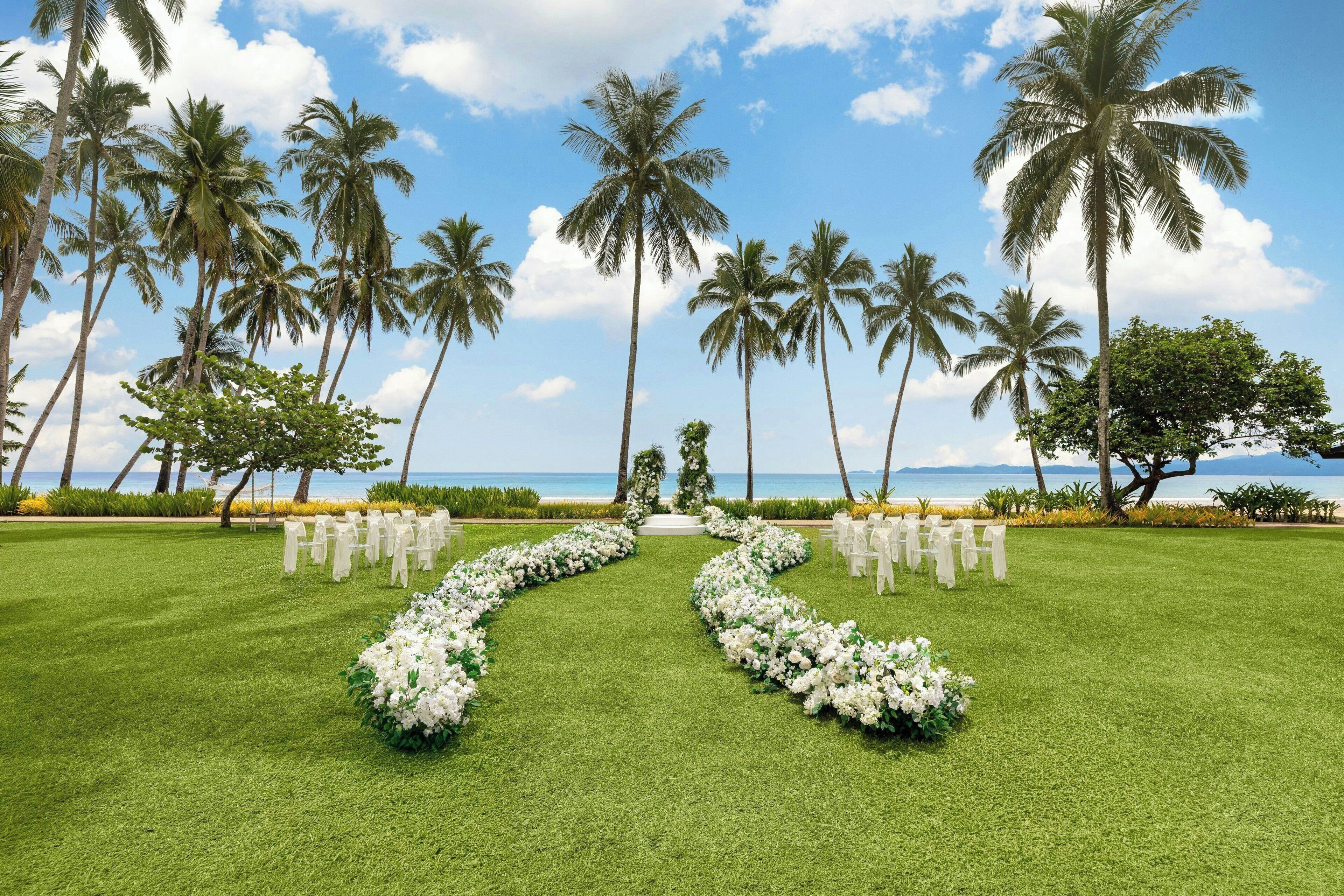 A wedding ceremony setup with a grassy aisle, white floral arrangements, and chairs with white covers, overlooking a beach with palm trees and the ocean.