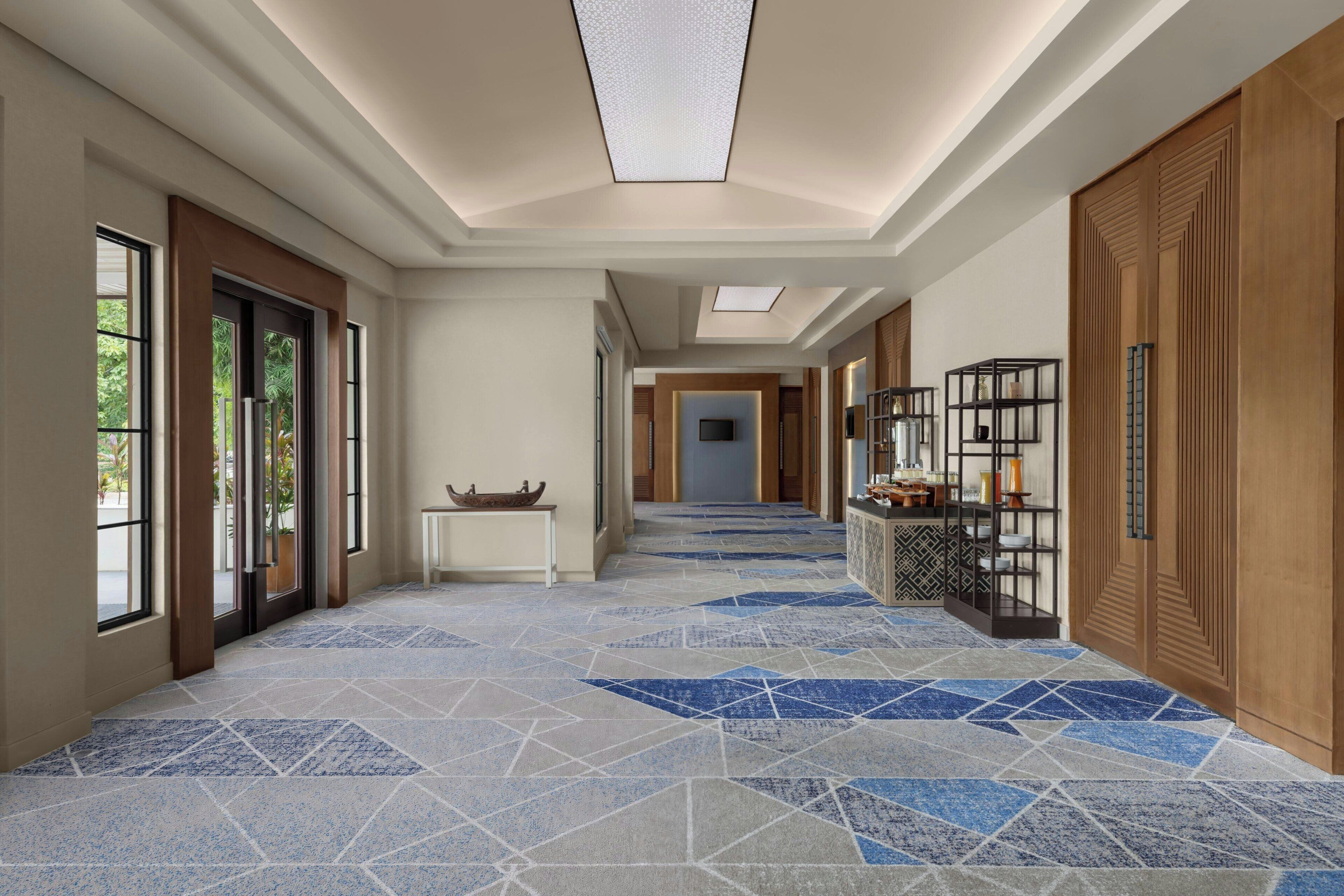 Pasillo de hotel con piso estampado, puertas de madera y una zona de recepción con mostrador y estanterías.