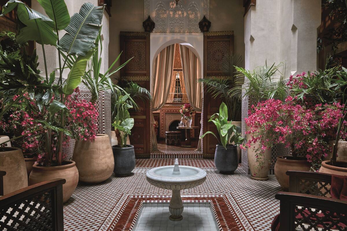 Un patio con una fuente, plantas en macetas y vista a una habitación con piso estampado y un arco decorativo.
