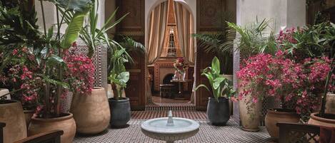 A courtyard with a fountain, potted plants, and a view of a room with a patterned floor and a decorative archway.
