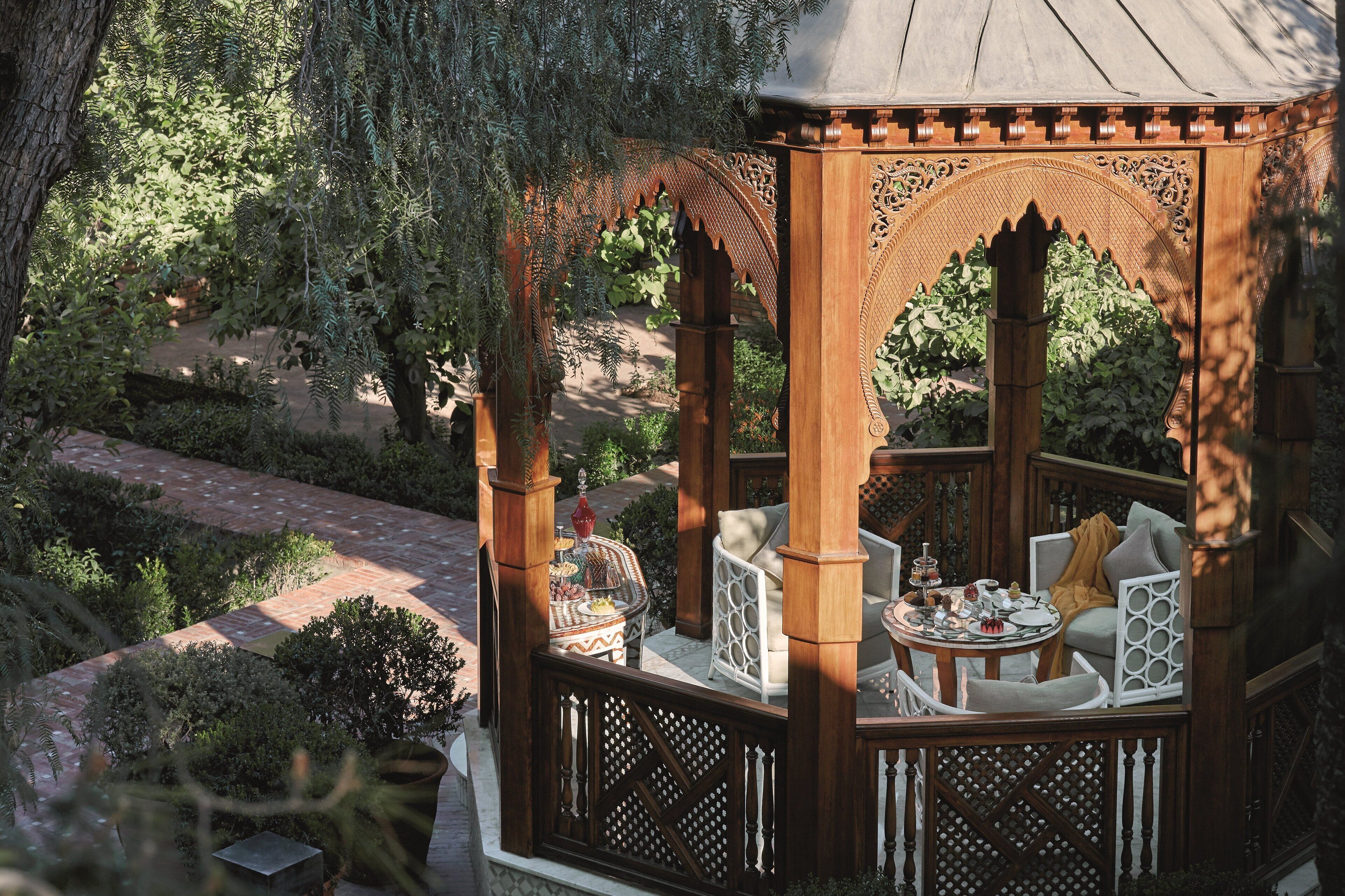 Un gazebo in legno con elaborate decorazioni intagliate, arredato con sedie bianche e un tavolo rotondo, immerso nel verde.