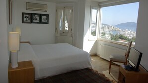 Ein Hotelzimmer mit einem Bett, einem Sessel, einem Schreibtisch mit Computer, einem Fenster mit Blick auf einen Yachthafen und die Stadtlandschaft sowie einem Heizkörper.