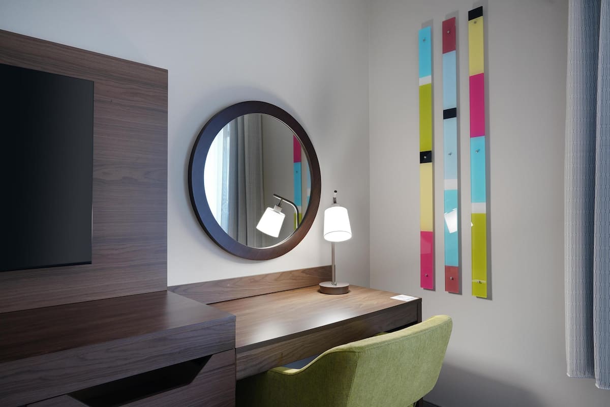 Moderni hotellihuone, jossa on puinen meikkipöytä, pyöreä peili, yöpöytälamppu, vihreä tuoli ja värikkäitä taideteoksia seinällä.