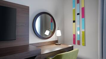 A modern hotel room with a wooden vanity, a round mirror, a bedside lamp, a green chair, and colorful artwork on the wall.