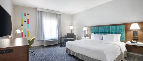 A hotel room with a large bed, a desk with a flat-screen TV, a chair, a small table, and a window with curtains.