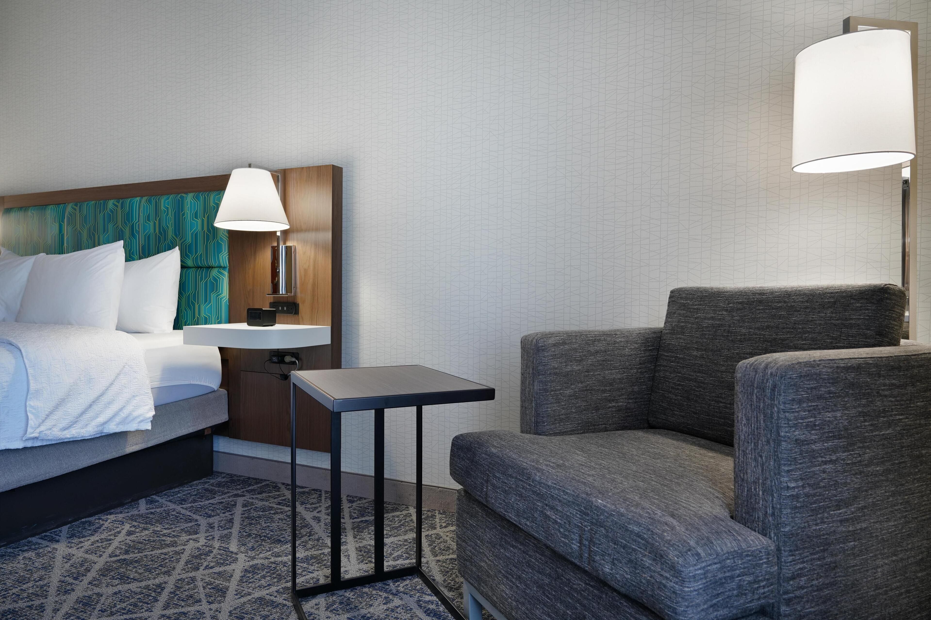 Een hotelkamer met een bed, een nachtkastje met een lamp, een klein tafeltje en een grijze fauteuil.