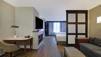 Ein Hotelzimmer mit Holzboden, einem Schreibtisch mit Lampe, einem Fernseher, einem Bett, einer Couch und einem Fenster mit Vorhängen.