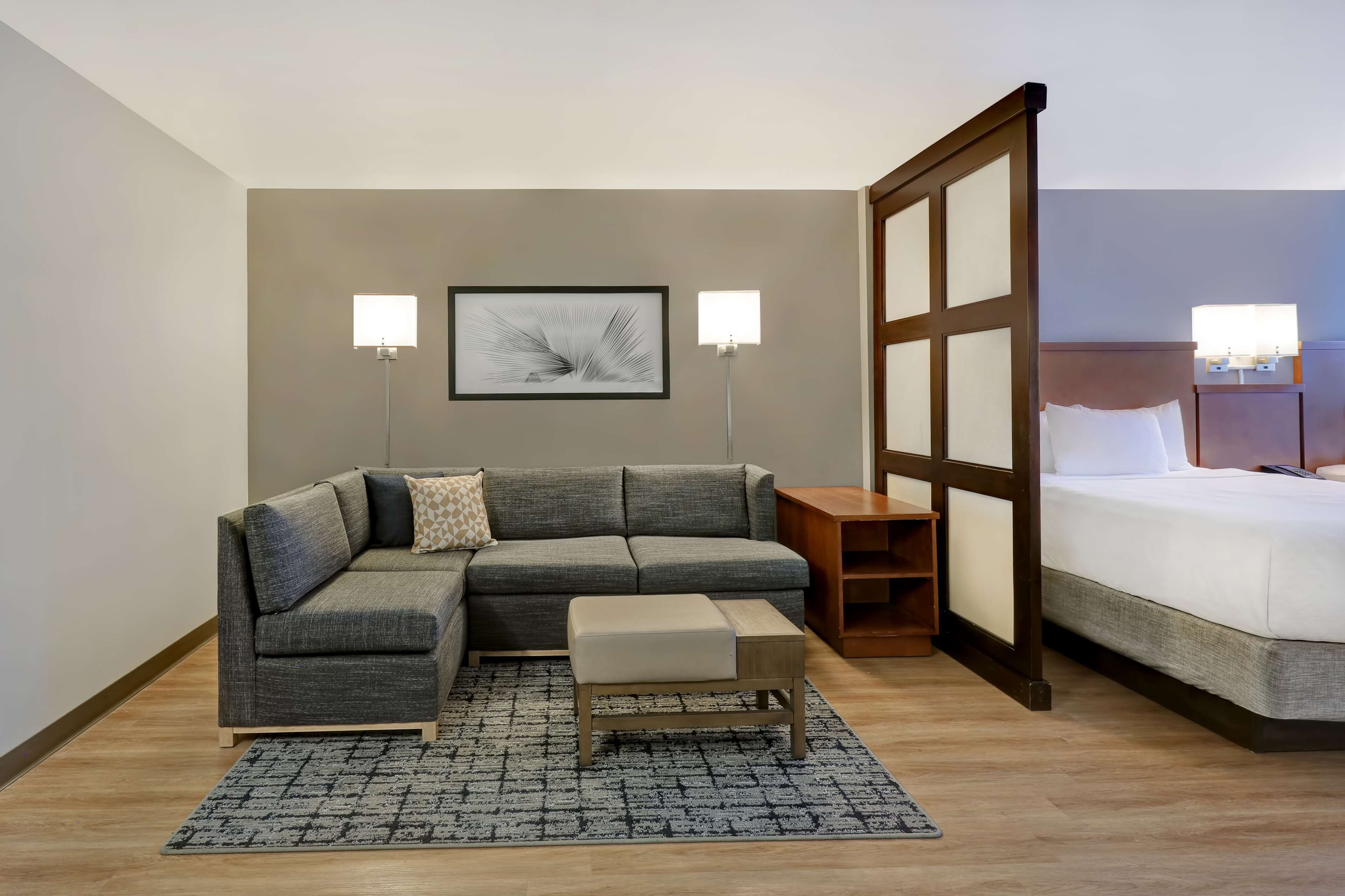 A modern hotel room with a grey sectional sofa, a small wooden side table, a footstool, a bed with white linens, and two wall-mounted lamps.