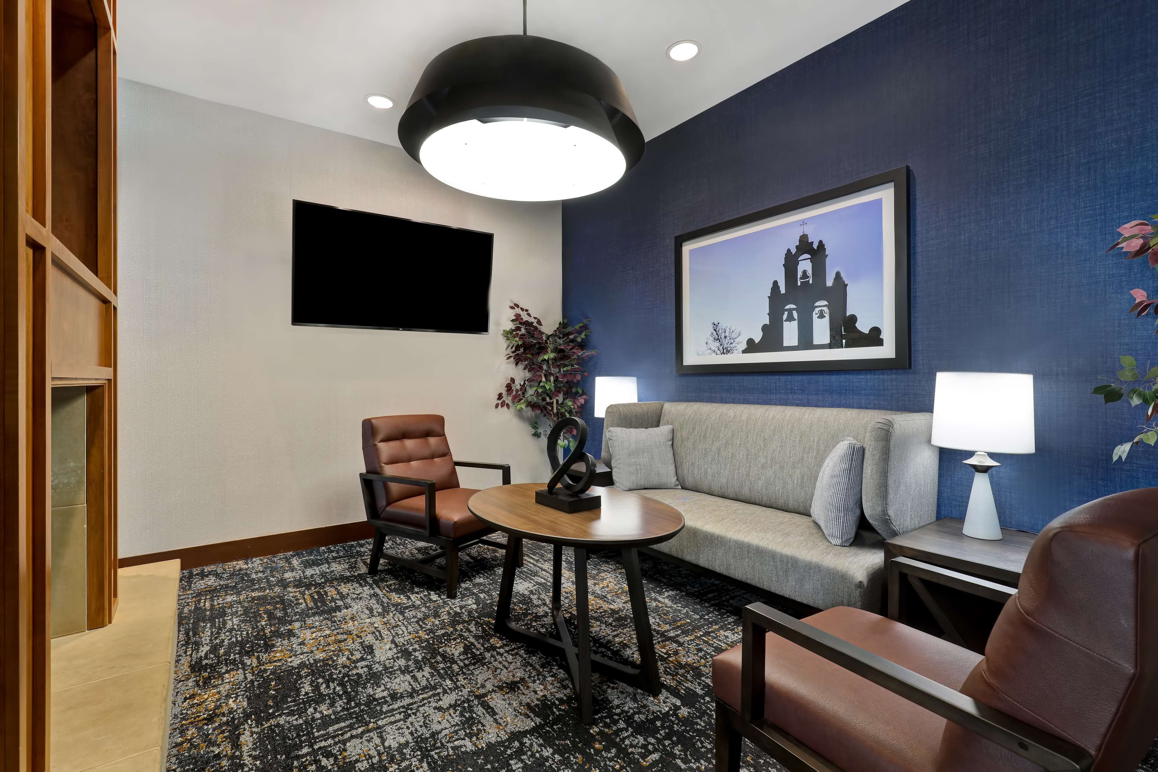 Una habitación de hotel moderna con una pared de acento azul oscuro, un cuadro de una iglesia, un televisor de pantalla plana, un sofá, dos sillones, una mesita redonda y una lámpara de pie.