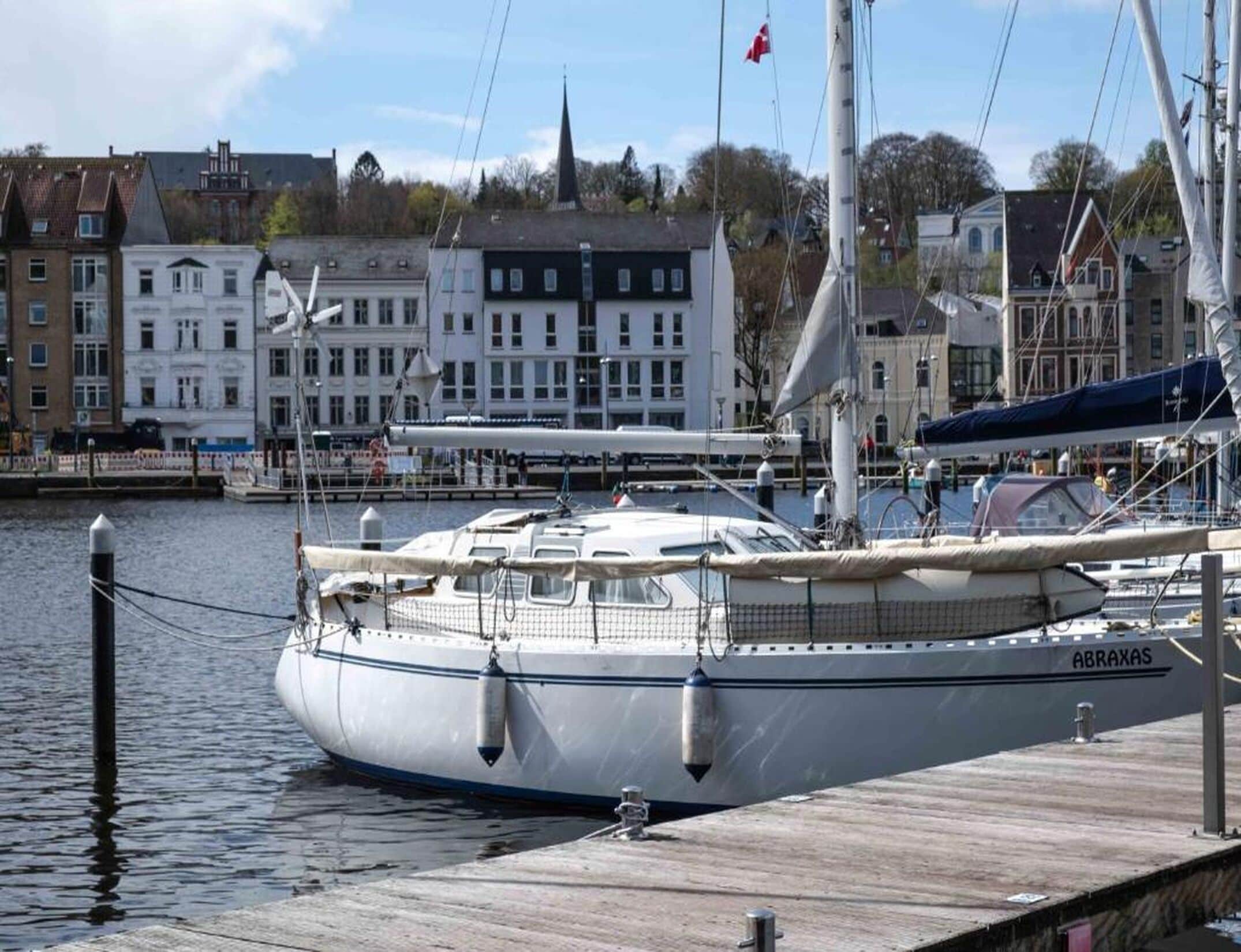 Un voilier est amarré dans une marina, avec des bâtiments en arrière-plan.