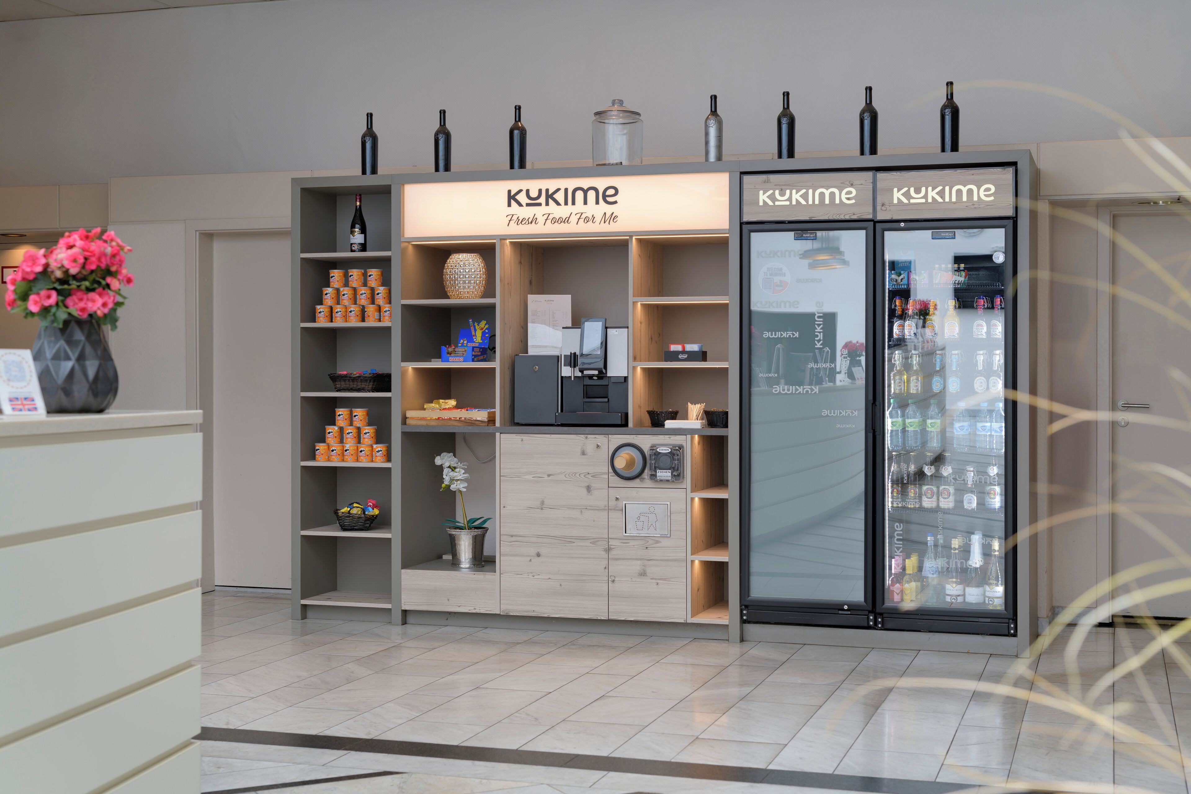 Un quiosco con un letrero que dice "KUKIME Fresh Food For Me", con una máquina de café, estantes con bocadillos y un refrigerador lleno de bebidas.
