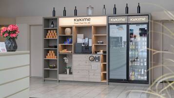 Un quiosco con un letrero que dice "KUKIME Fresh Food For Me", con una máquina de café, estantes con bocadillos y un refrigerador lleno de bebidas.