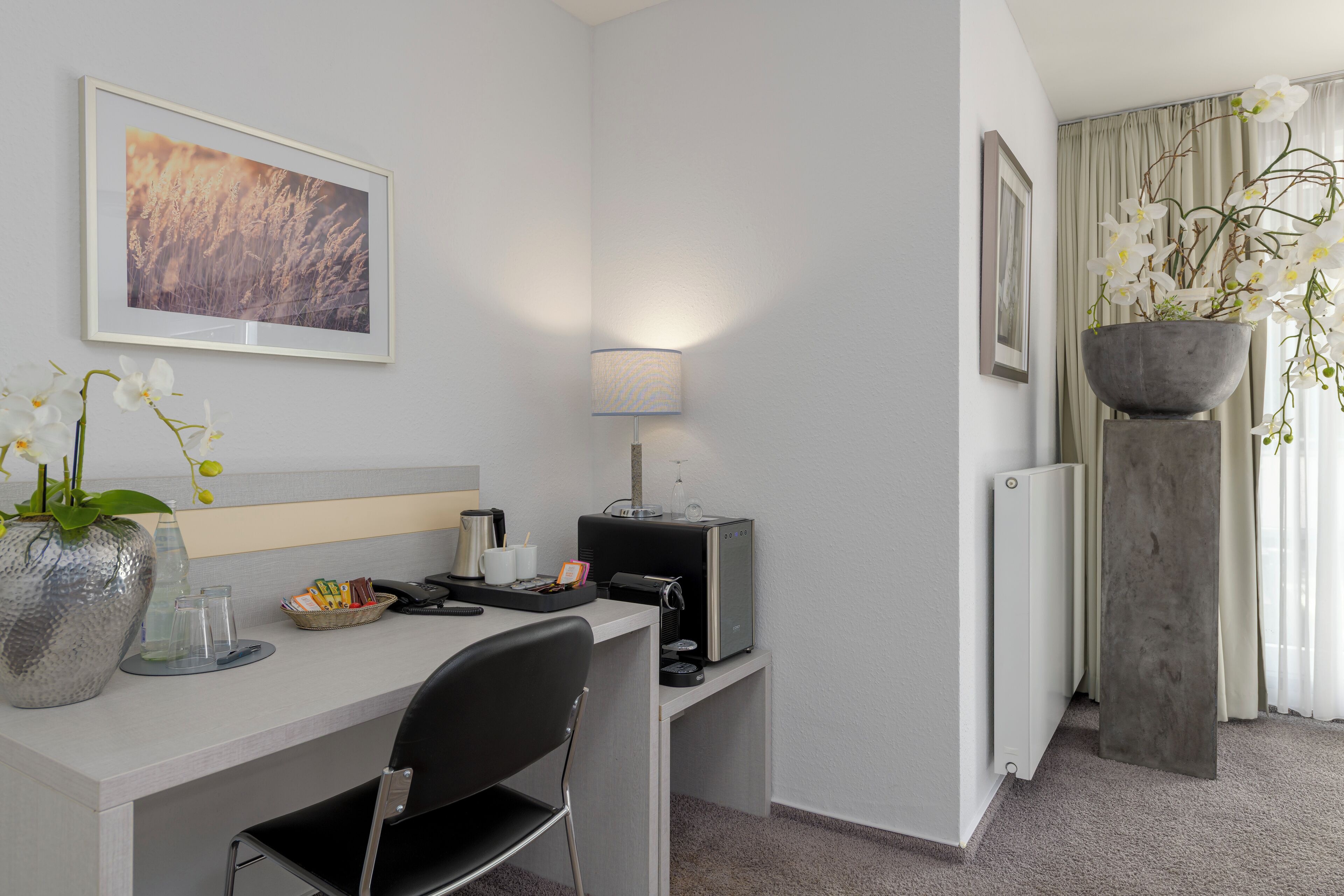 Hotellihuone, jossa on työpöytä, tuoli, kahvinkeitin, lamppu ja suuri ikkuna, jossa on verhot.