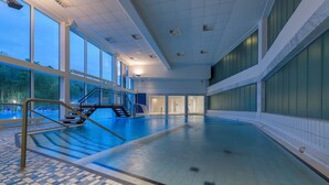 A large indoor swimming pool with clear glass walls, a tiled floor, and a staircase leading to a second level. - Sunday Hotel des Nordens (Harrislee)