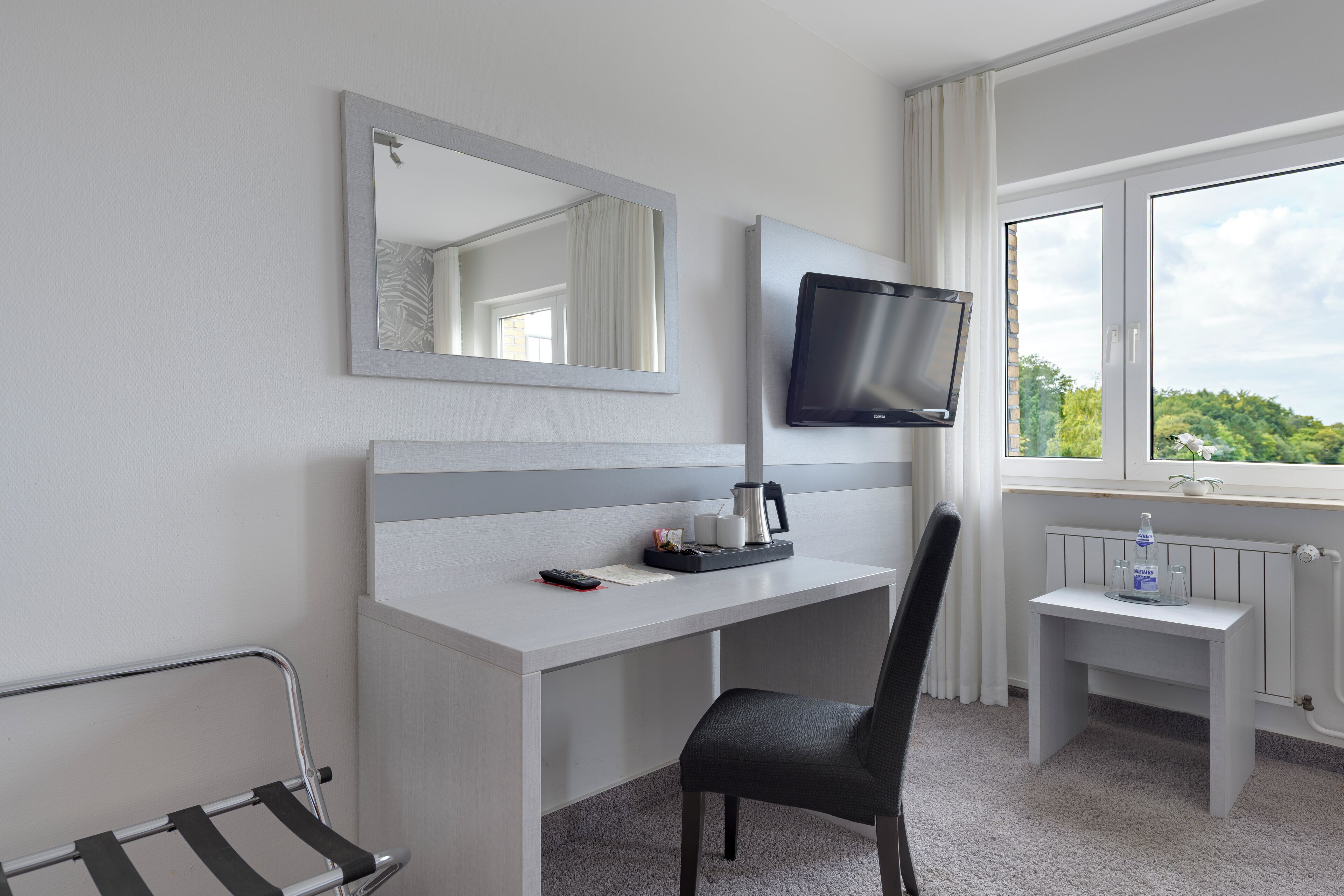 Habitación de hotel con un escritorio blanco, una silla negra, un televisor, una mesita con una botella y una ventana con vistas a zonas verdes.