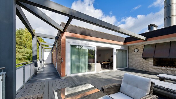 A covered outdoor deck with wicker furniture, a glass door leading to a patio, and a brick wall. - Sunday Hotel des Nordens (Harrislee)