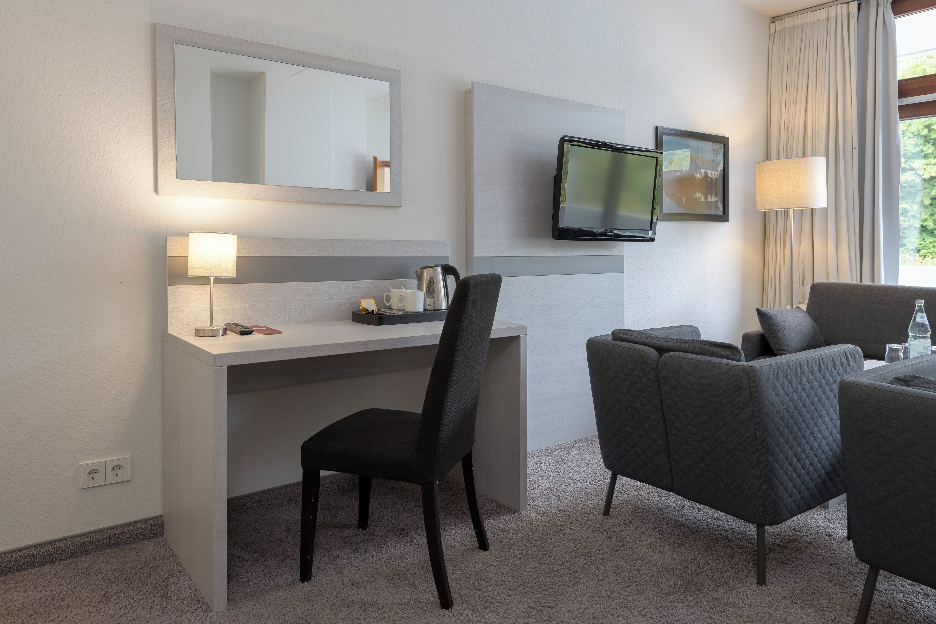 A hotel room with a desk, chair, sofa, TV, and a window with curtains.
