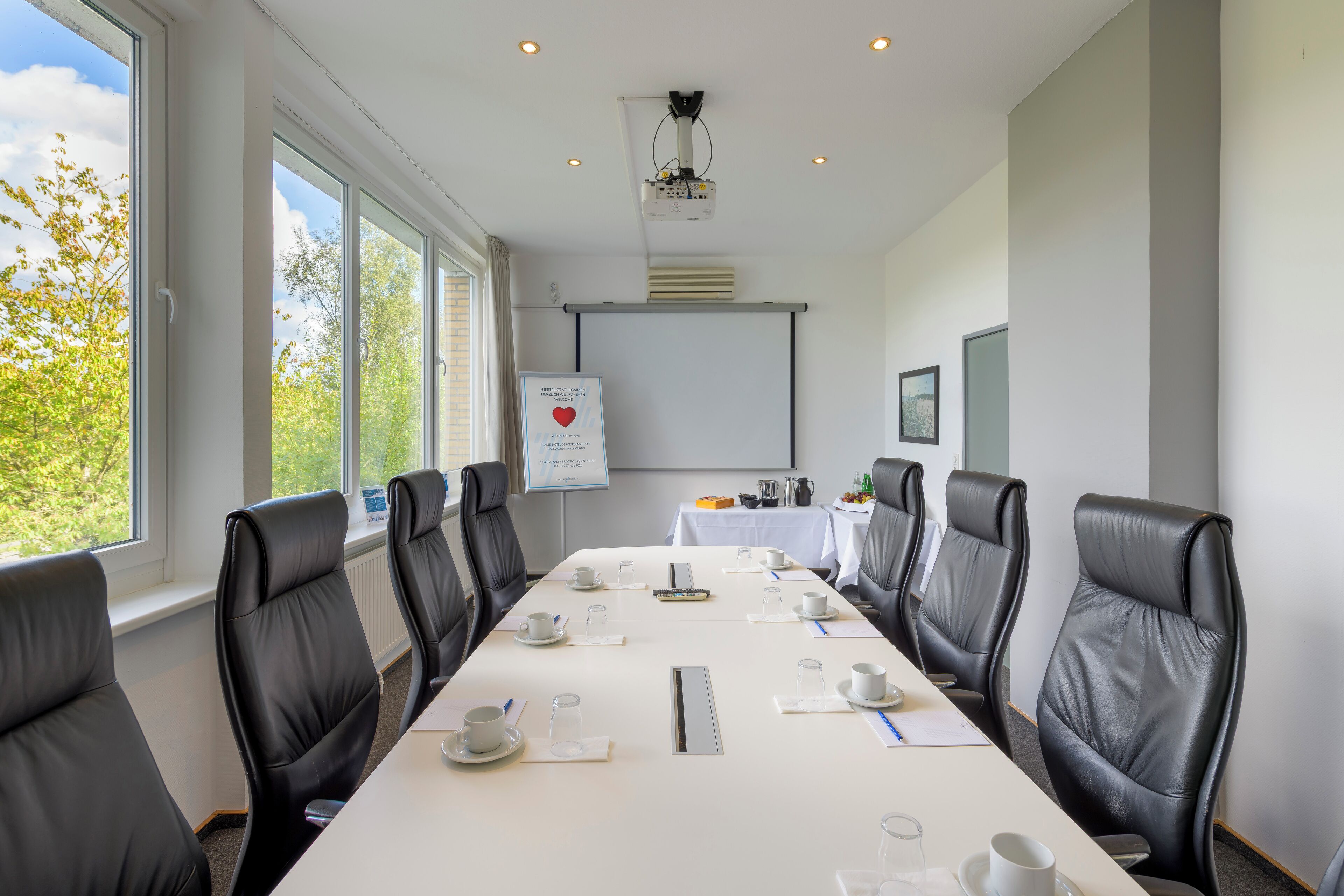 A conference room with a long table, black leather chairs, white tablecloths, and a projector screen.