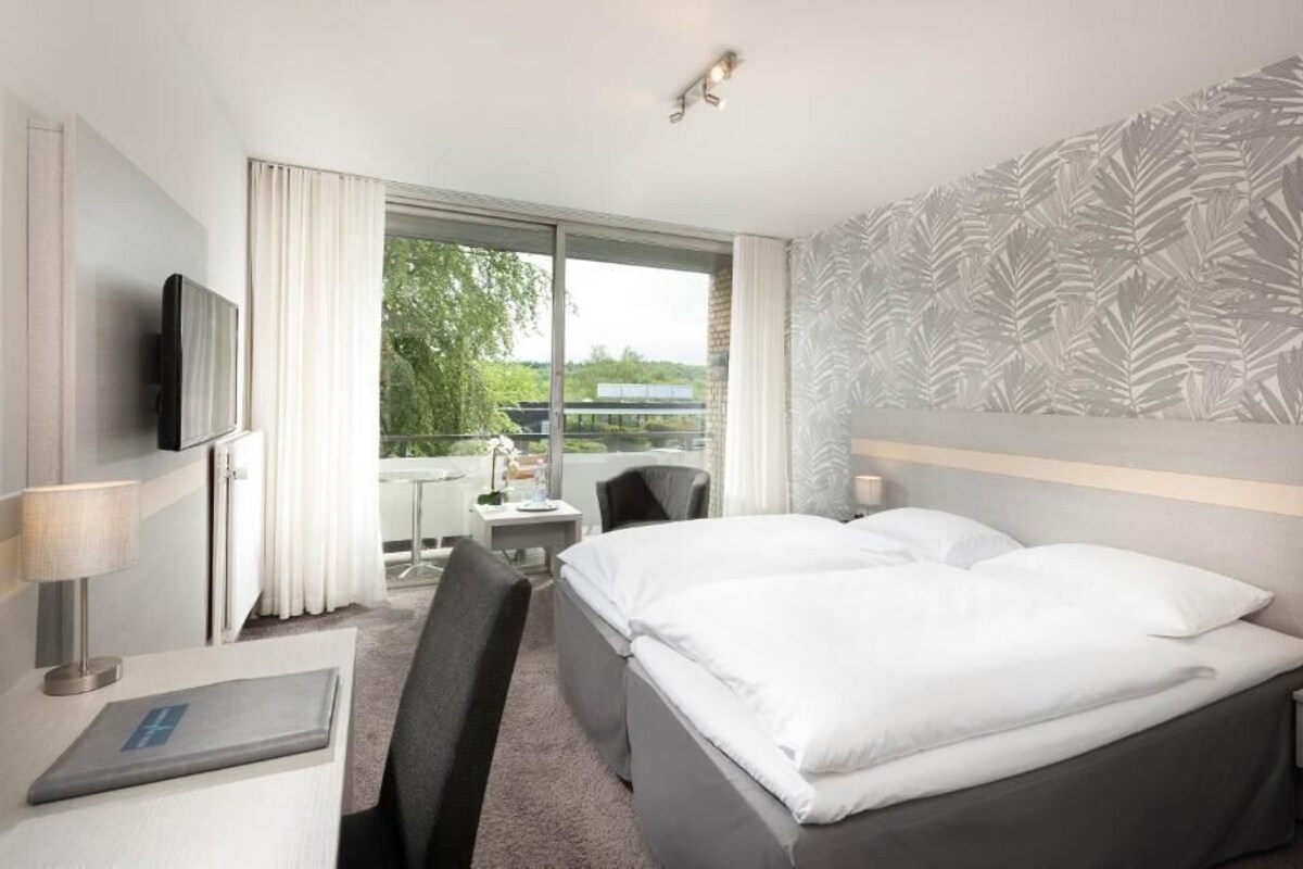 A hotel room with a bed, a desk, a chair, a television, and a window with a view of greenery.