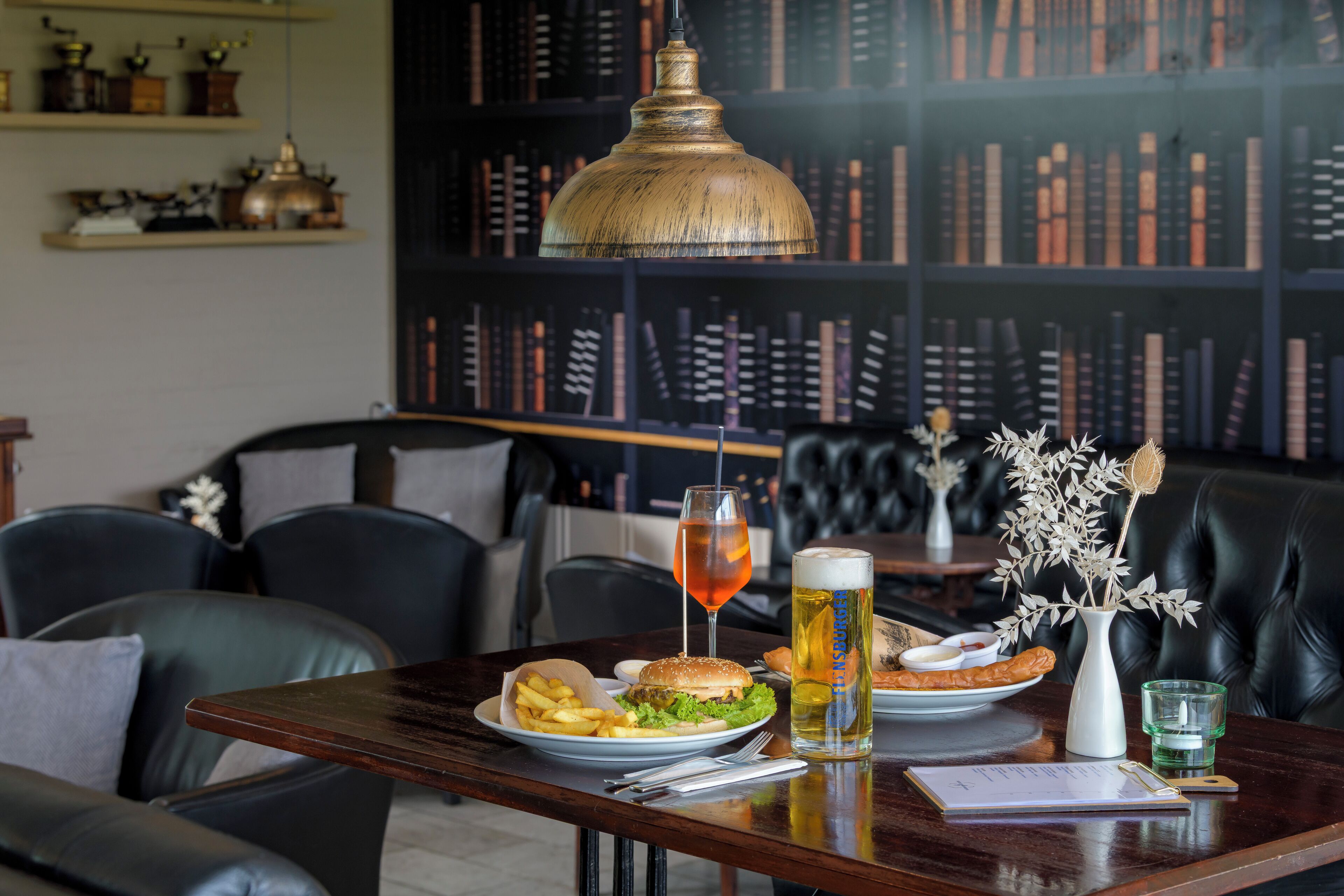 A dining table with a burger, fries, and a drink, set against a backdrop of bookshelves and a hanging lamp.