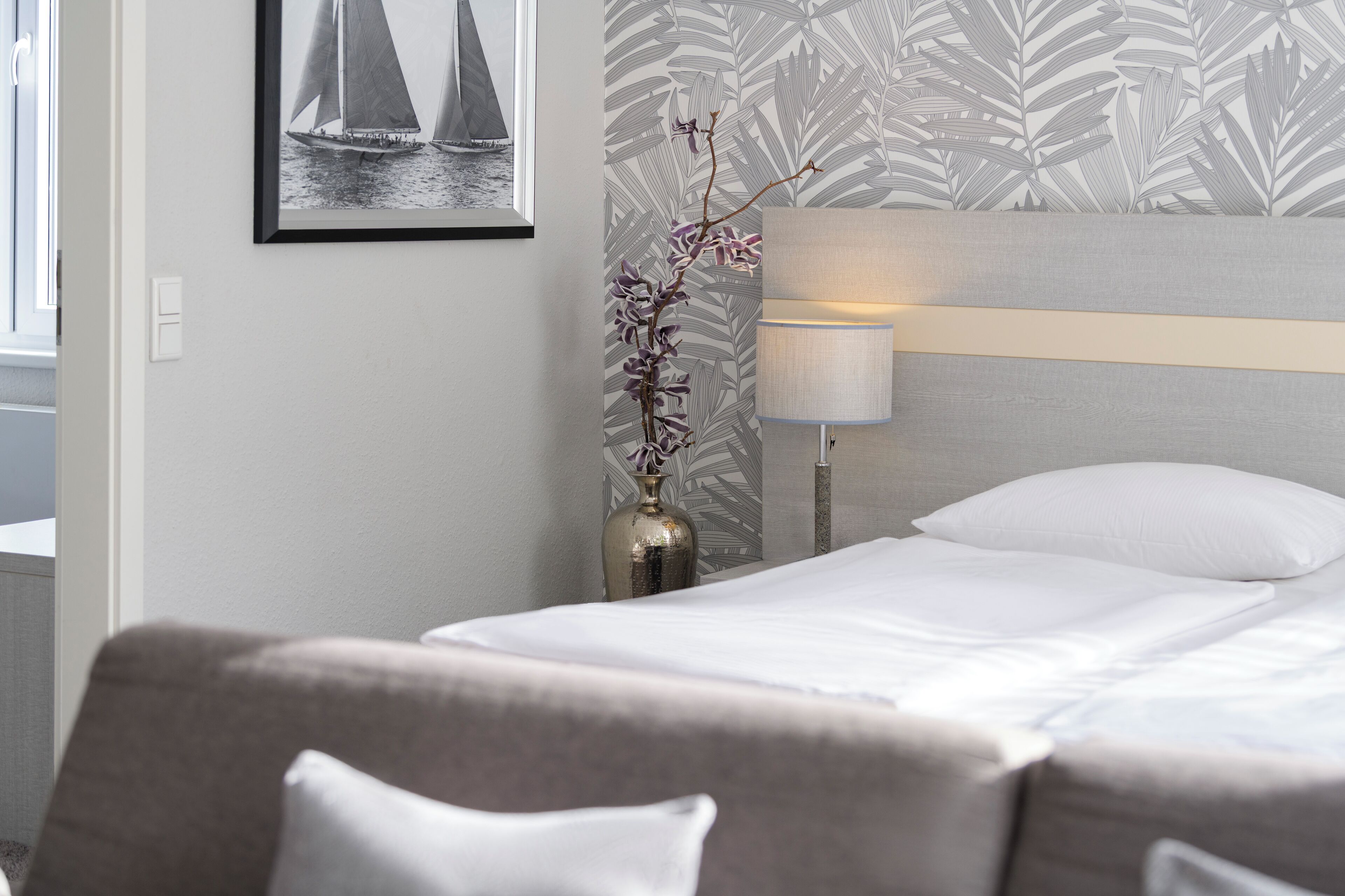 A neatly arranged hotel room with a bed, a nightstand with a lamp, a vase with flowers, and a framed picture on the wall.