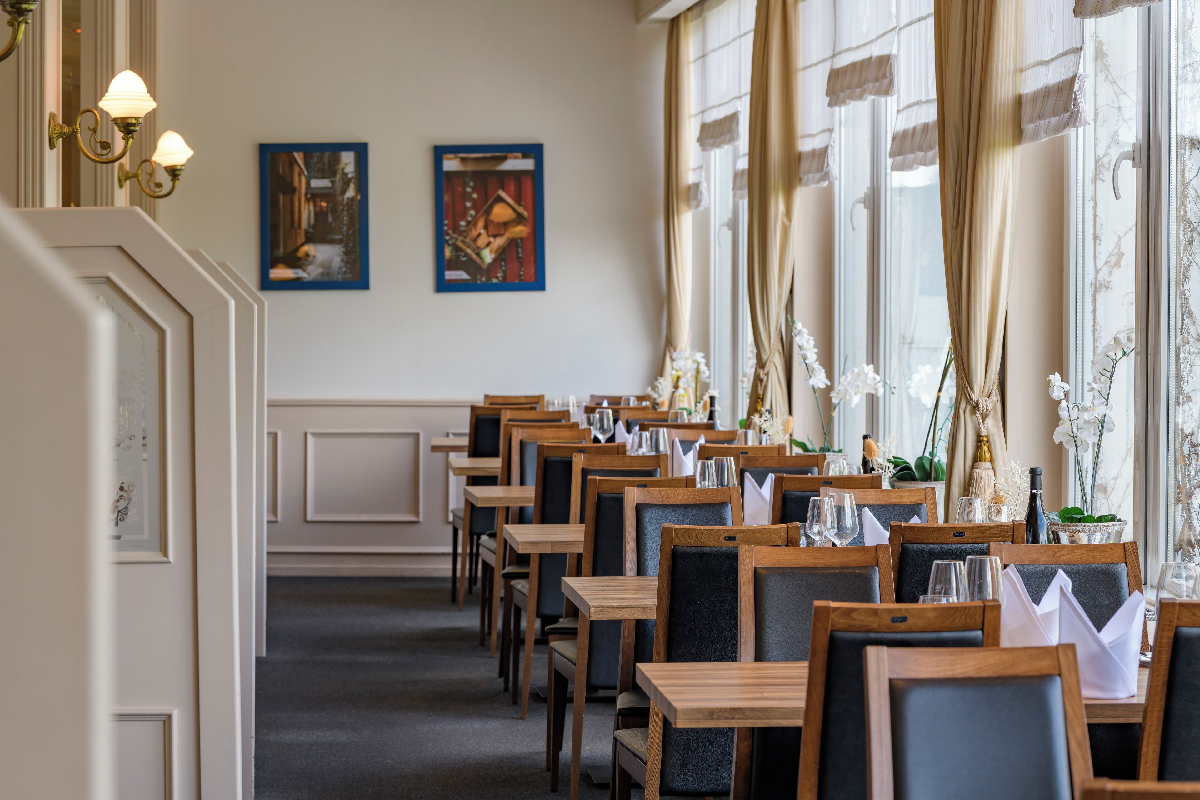 A restaurant with wooden tables and chairs, framed artwork on the walls, and large windows with curtains.
