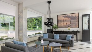 A modern living room with a grey sofa, two side tables, and a large window offering a view of greenery. - Sunday Hotel des Nordens (Harrislee)