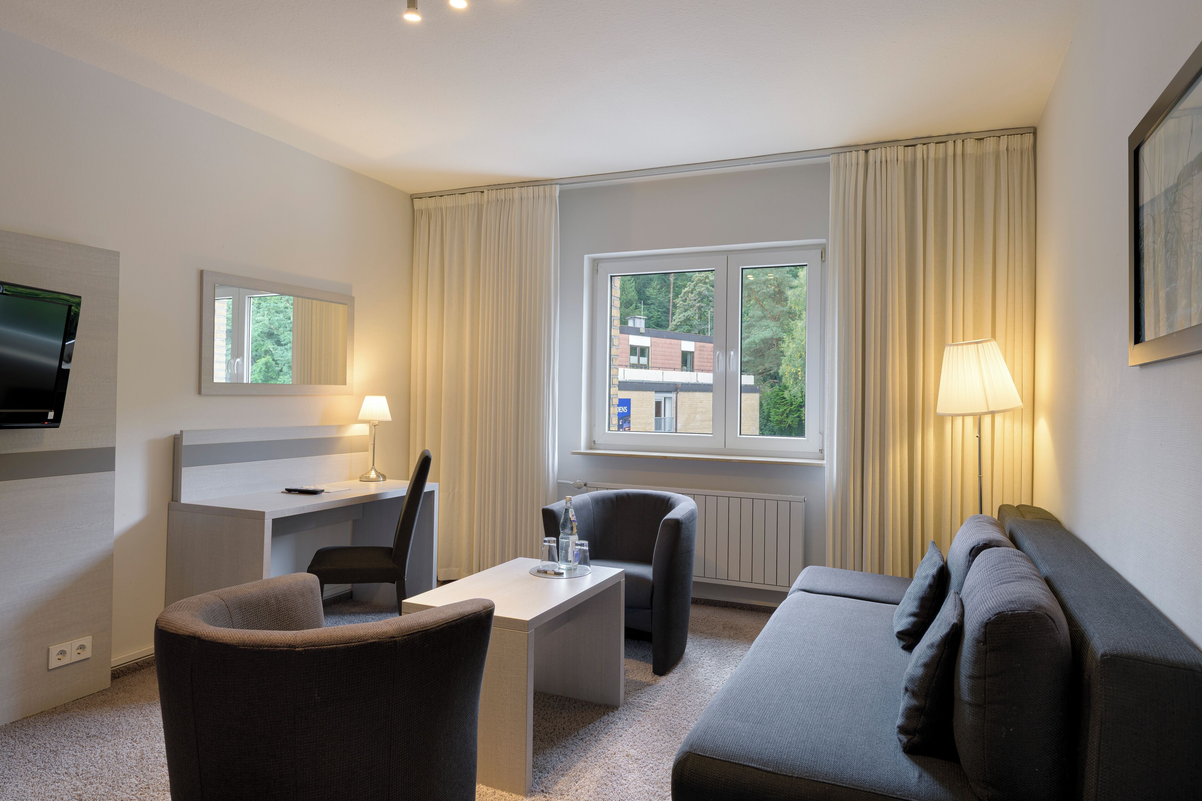 Habitación de hotel con escritorio, dos sillas, sofá, televisión y ventana con cortinas.