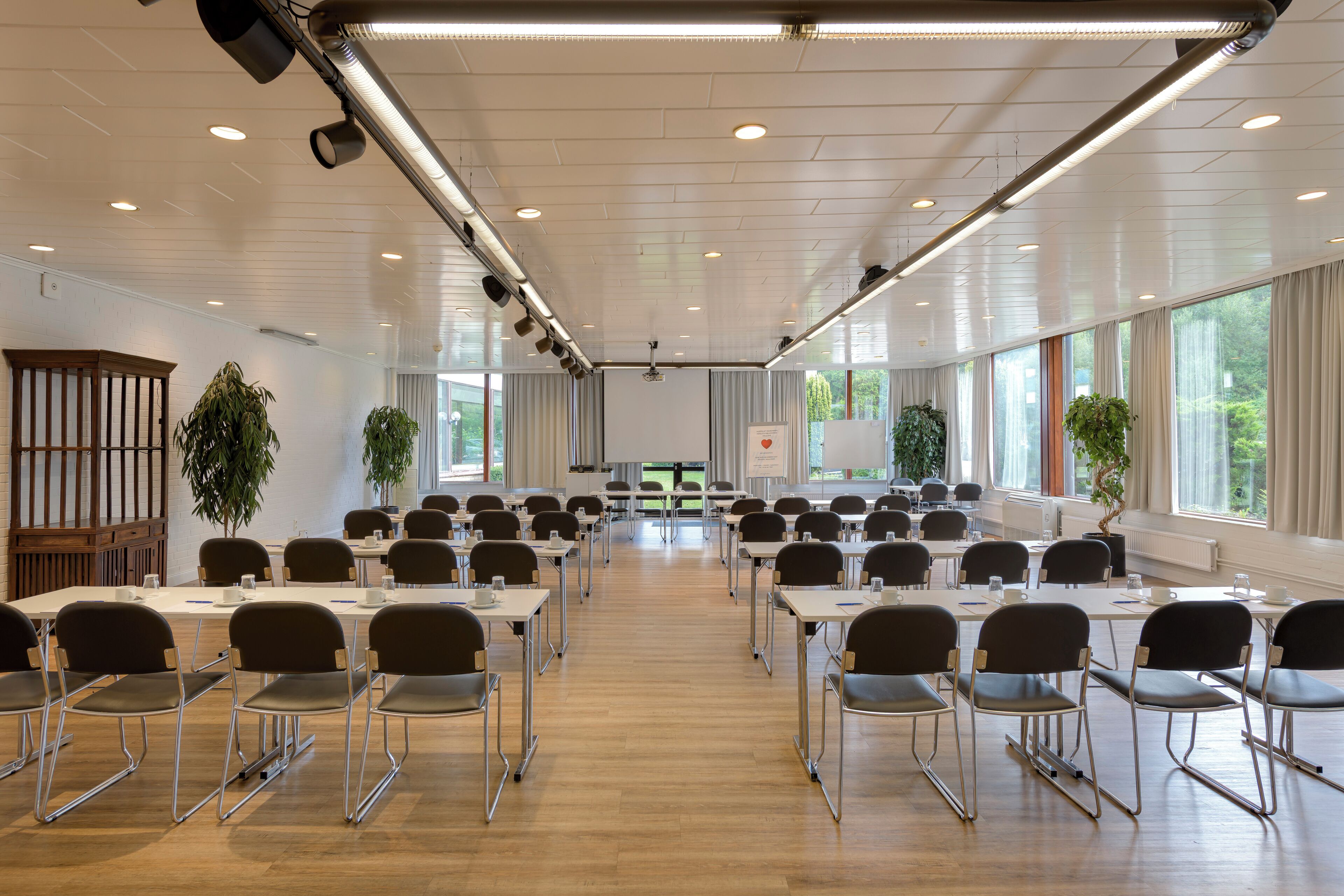 Uma sala de conferências com mesas e cadeiras dispostas em fileiras, janelas amplas e luminárias no teto.