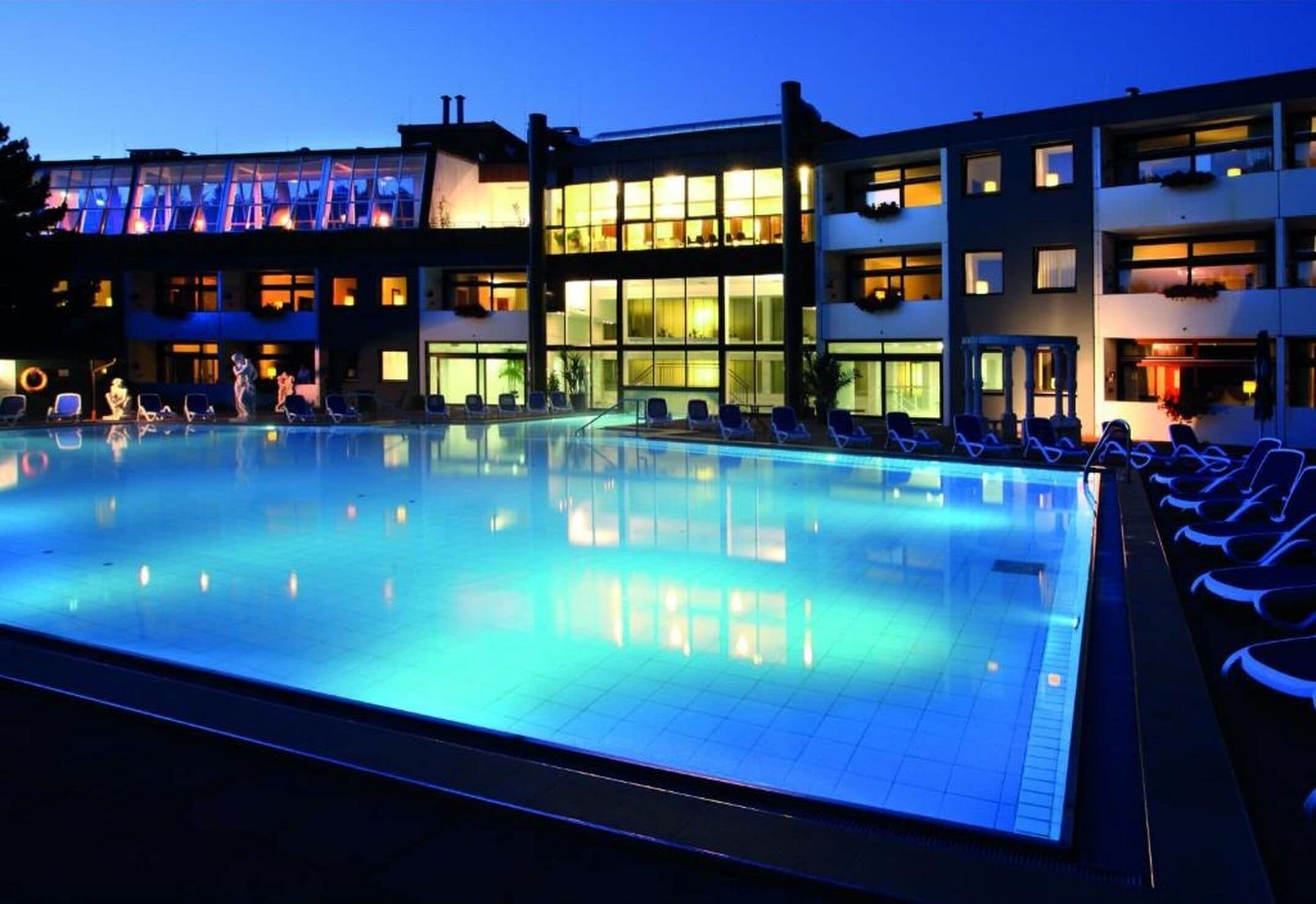 Una piscina con sillas de descanso, un edificio de varios pisos y amplias ventanas, y un cielo despejado.