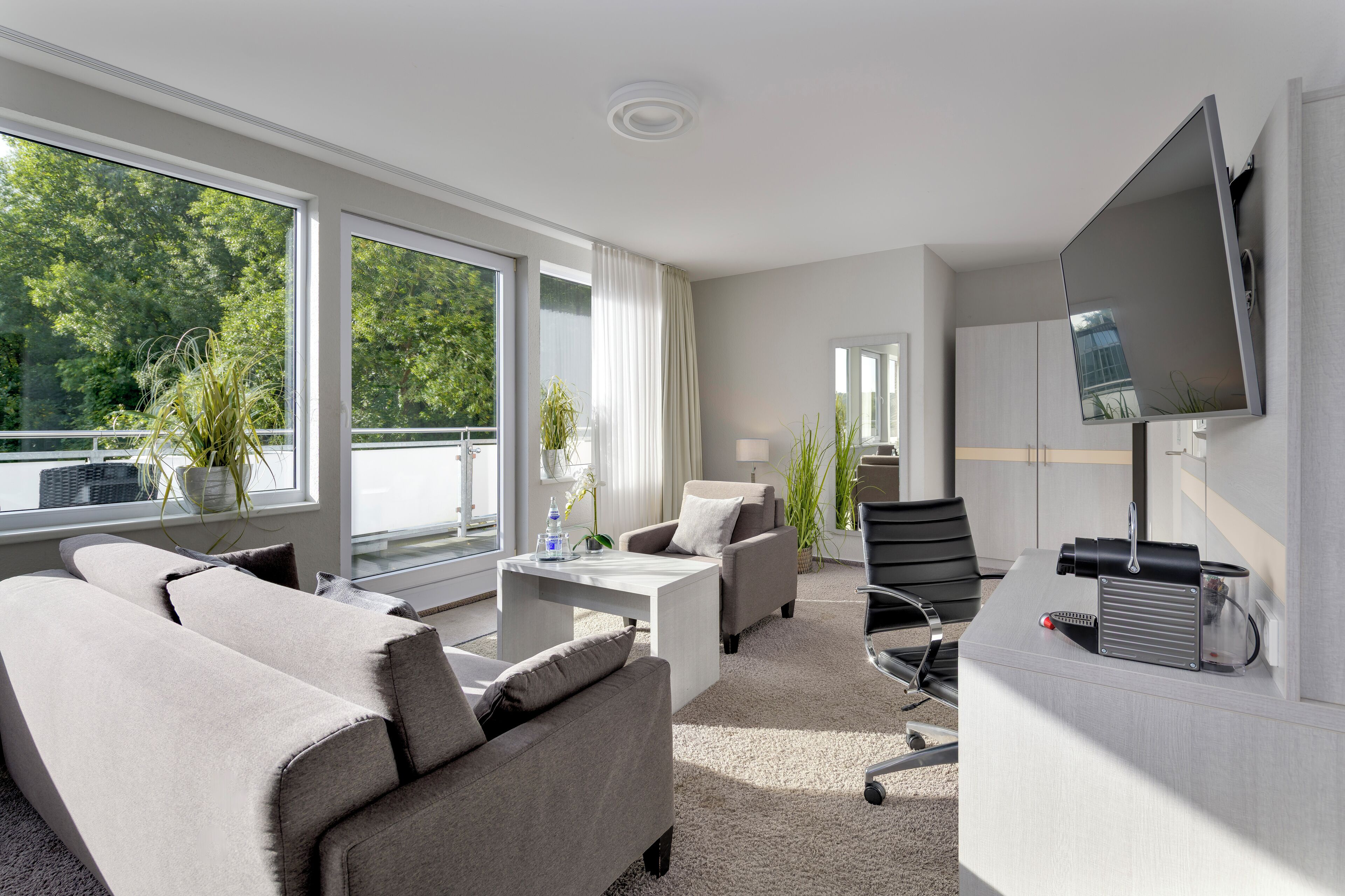 A modern hotel room with a sofa, coffee table, and TV. Large windows offer a view of trees and a balcony.