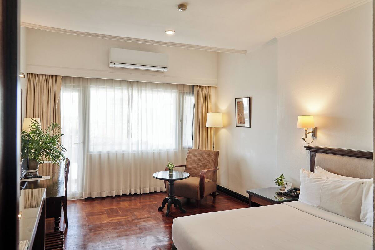 Een hotelkamer met een bed, een stoel, een kleine tafel met een plant, een lamp en een airconditioning.