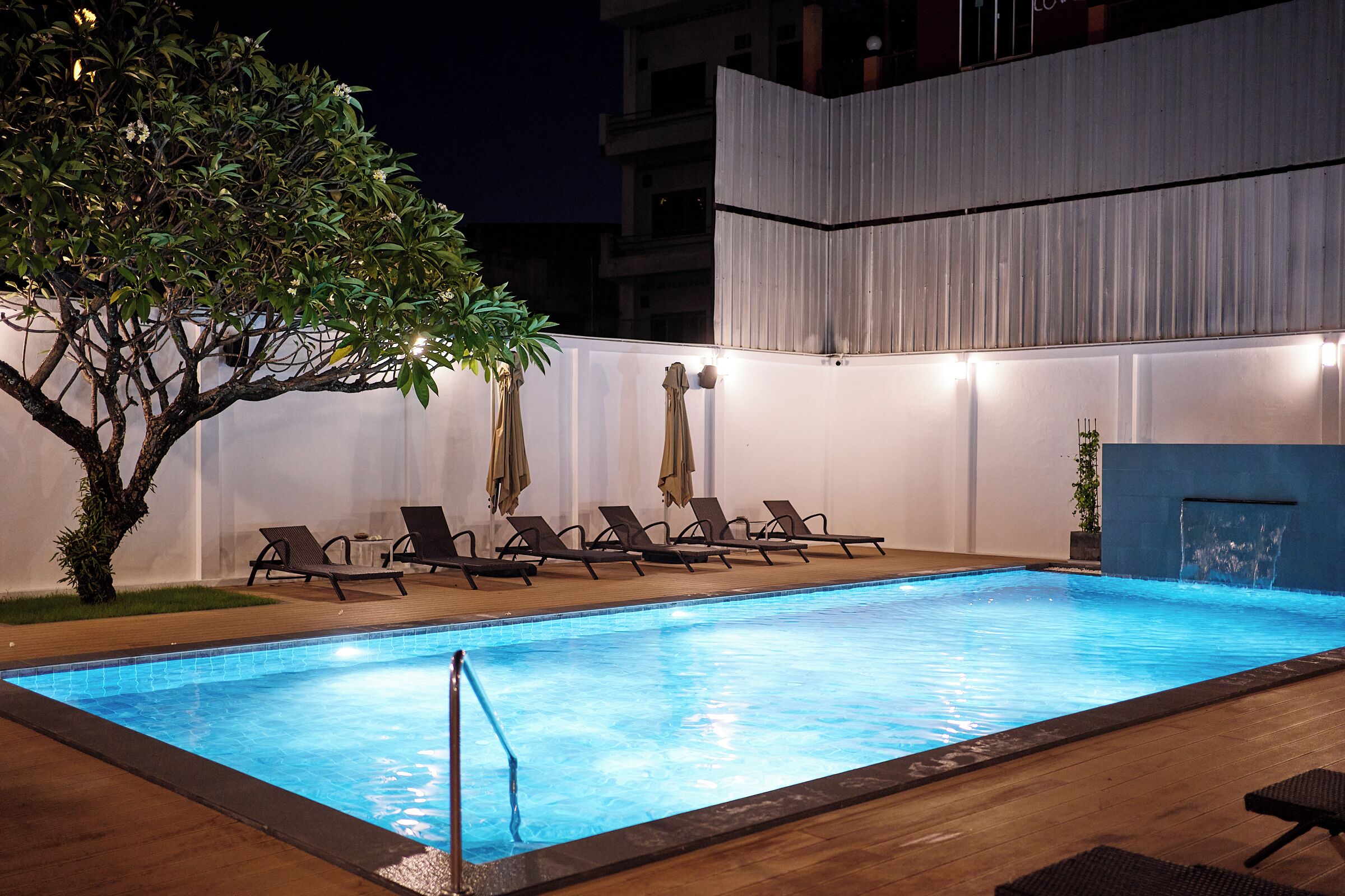 Una piscina interior con sillones de descanso y sombrillas, rodeada por un muro blanco y un árbol.