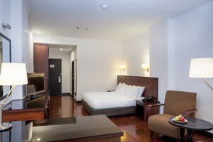 A hotel room with a bed, a desk, a chair, a TV, and a small table with fruit.