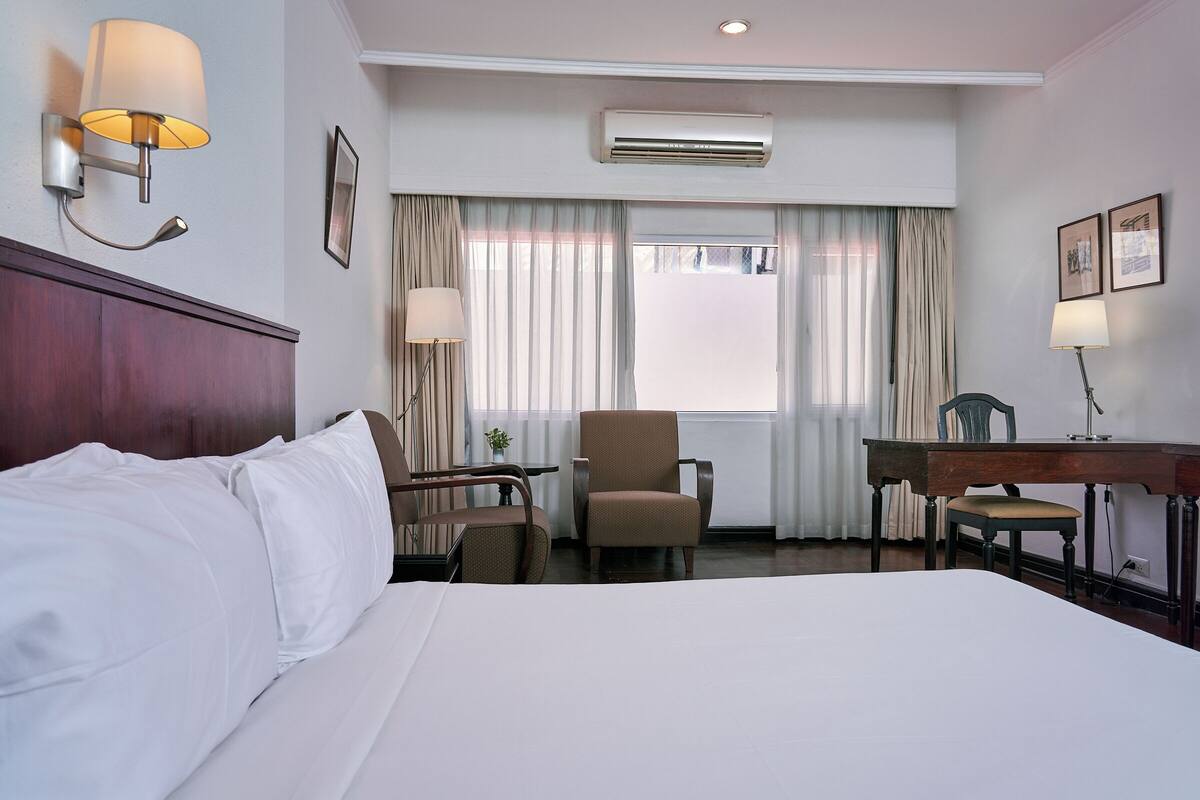 Een hotelkamer met een bed, twee stoelen, een bureau, een airconditioner en een raam met gordijnen.