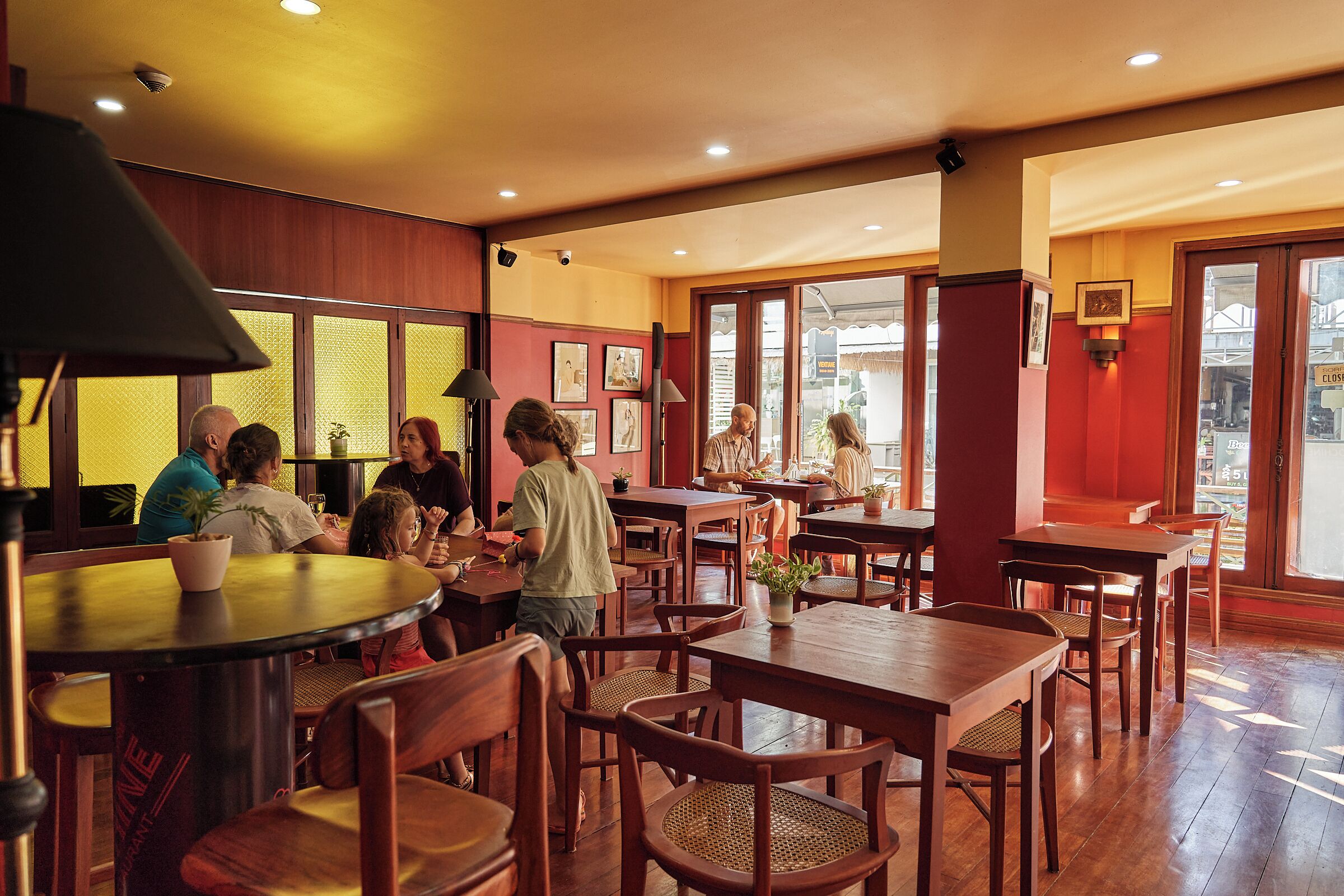 Un restaurant avec des tables et des chaises en bois, un coin bar et de grandes fenêtres laissant entrer la lumière naturelle.