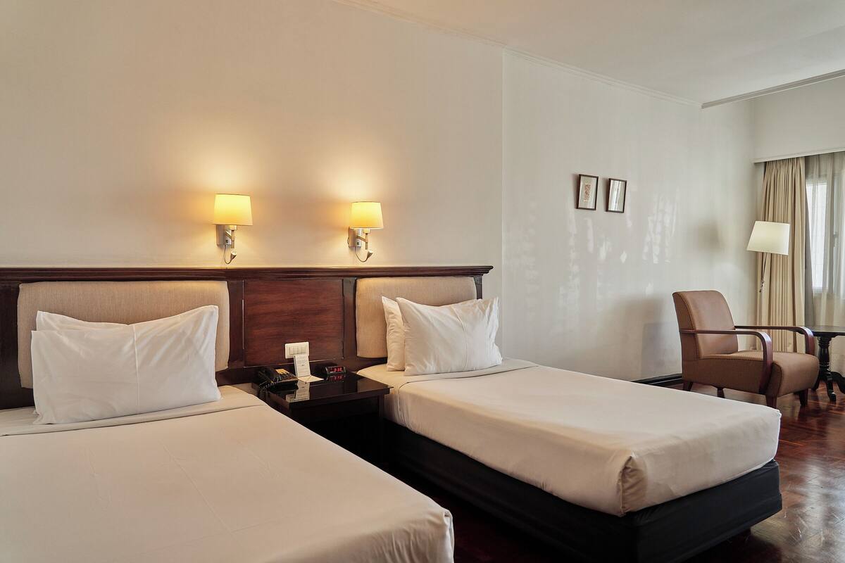 Een hotelkamer met twee bedden, bedlampen, een nachtkastje, een stoel en een raam met gordijnen.