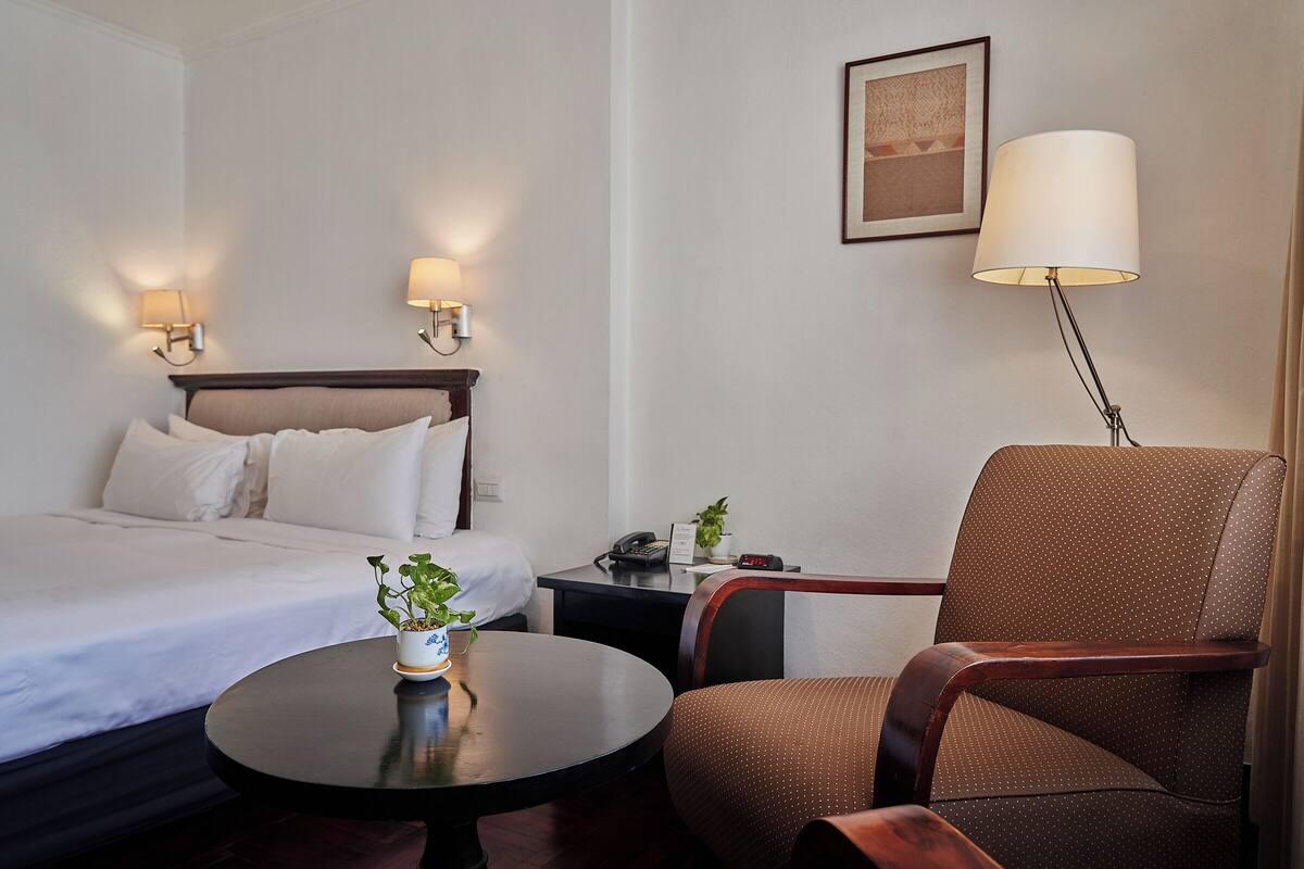 Een hotelkamer met een bed, een ronde tafel met een potplant, een bruine fauteuil, een bureau met een lamp en een lijst met een afbeelding aan de muur.